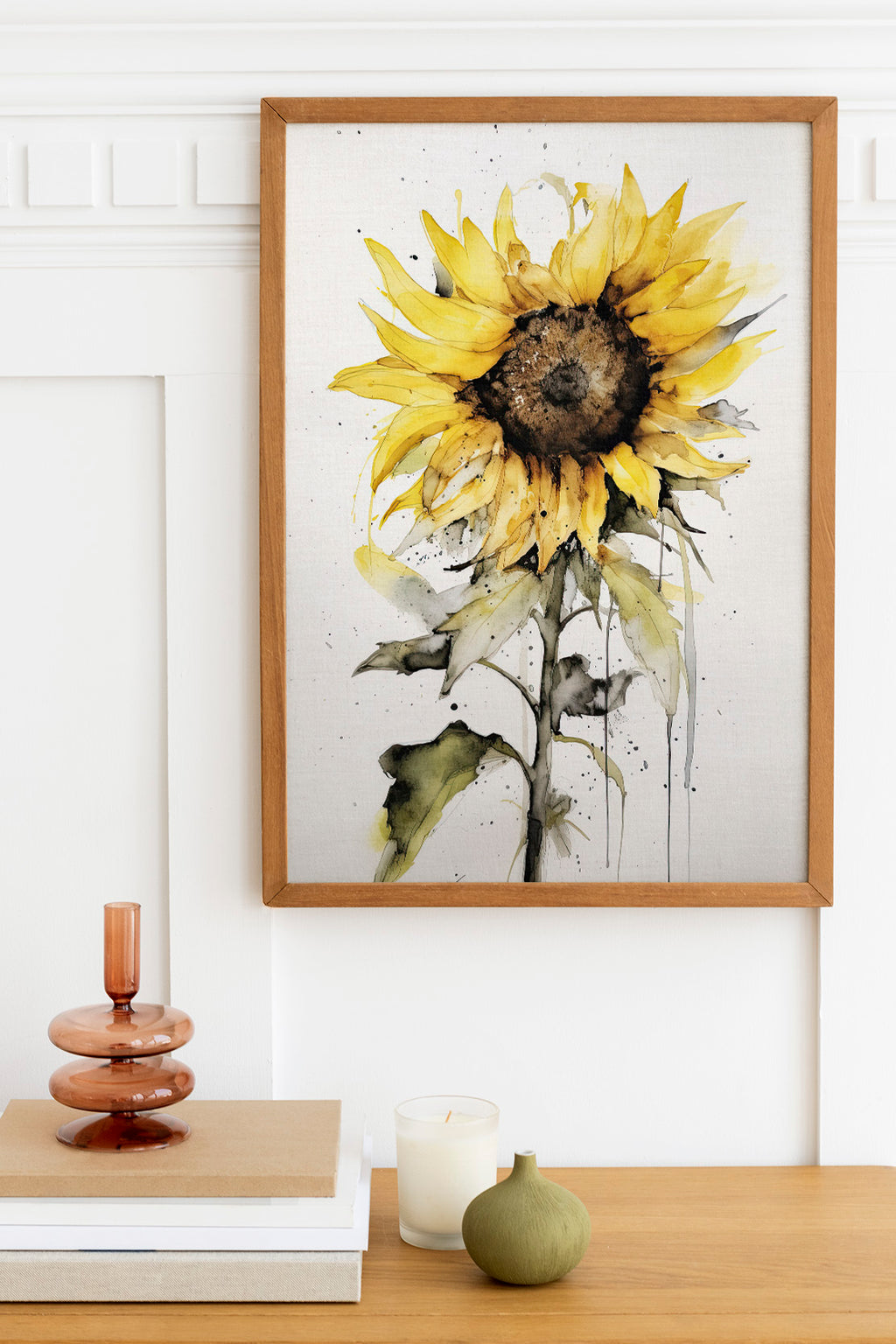 a framed watercolor painting of a yellow sunflower hanging on a wall above a wooden table with various decorative items, including a candle, a small green vase, and a stack of books.