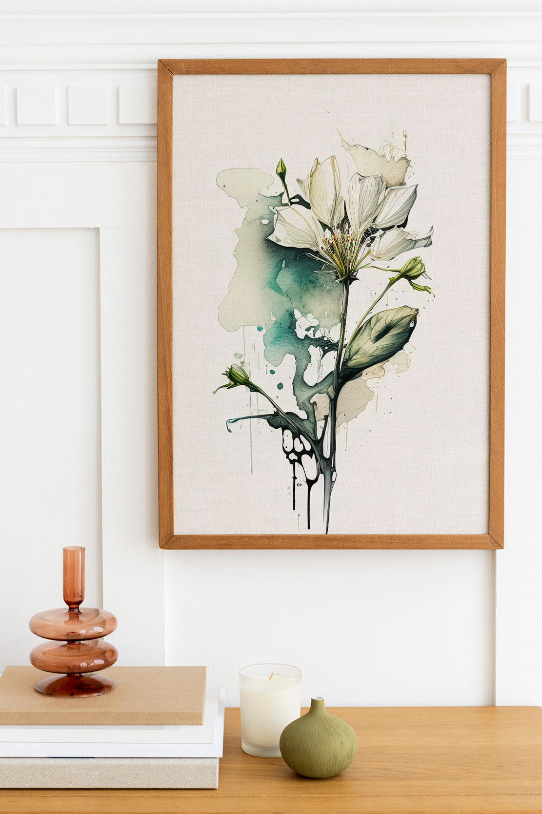 a framed watercolor painting of a white flower with green leaves, hanging on a white wall above a wooden table with various decorative items, including a stack of books, a small green vase, and a white candle.