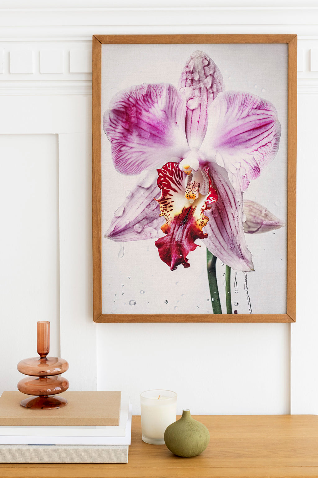 a framed photograph of a large purple orchid flower hanging on a wall above a wooden table. The table has a stack of books, a small green vase, and a white candle.