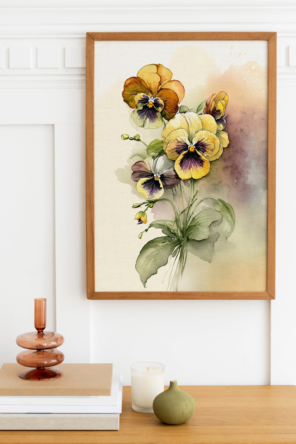 a framed painting of a bouquet of colorful flowers hanging on a wall above a wooden table. The table has a stack of books, a small green vase, and a white candle.