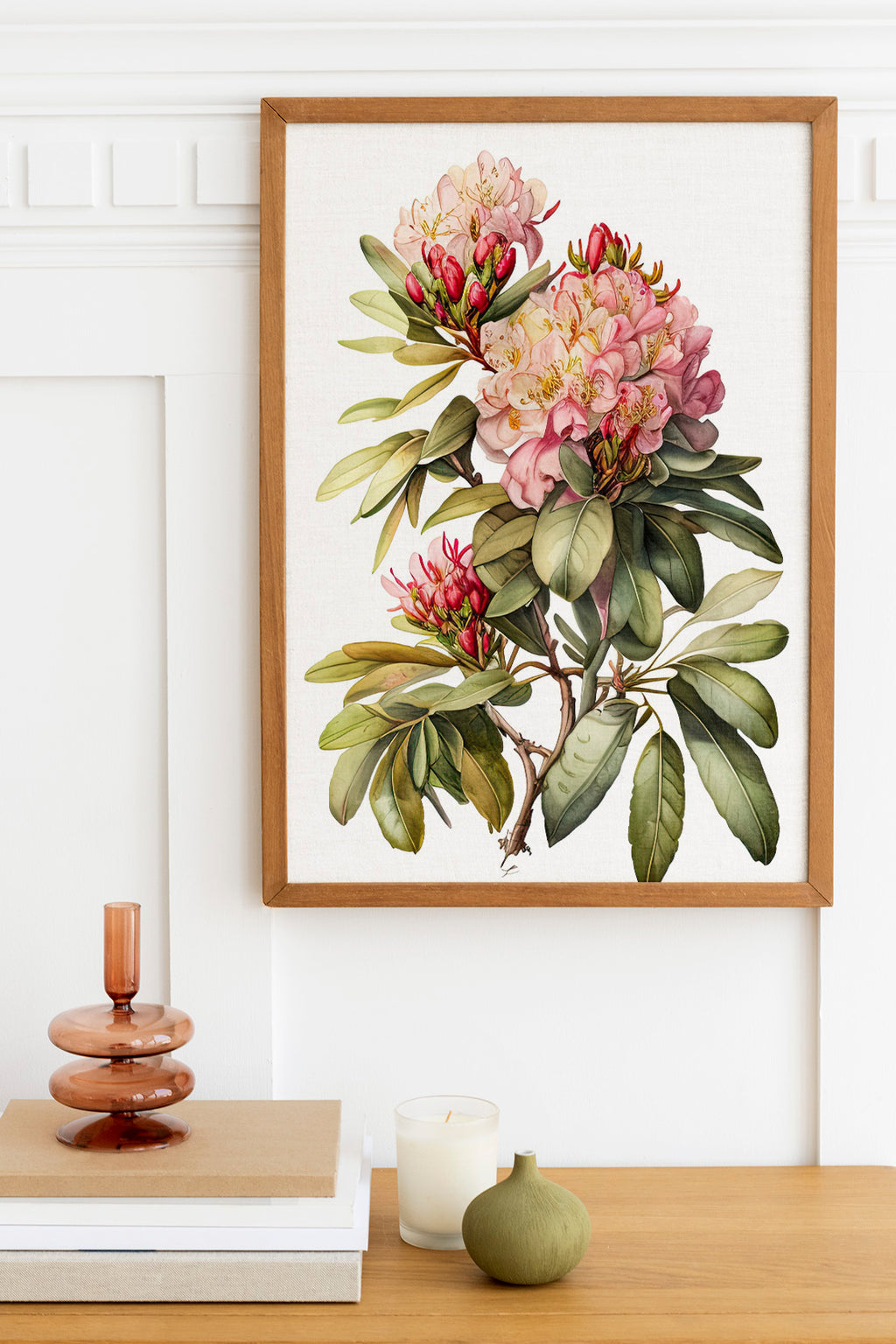 a framed botanical illustration of a pink and white flower on a white wall, with a wooden shelf below it holding a stack of books, a small green vase, and a white candle.
