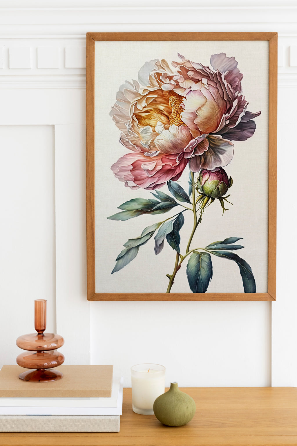 a framed painting of a large, colorful flower hanging on a wall above a wooden table with various decorative items, including a candle, a small green vase, and a stack of books.