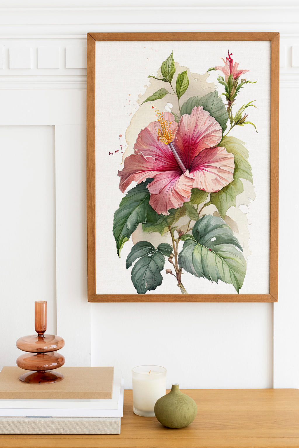 a framed painting of a large pink hibiscus flower with green leaves, hanging on a white wall above a wooden table. On the table, there is a stack of books, a small green vase, and a white candle.
