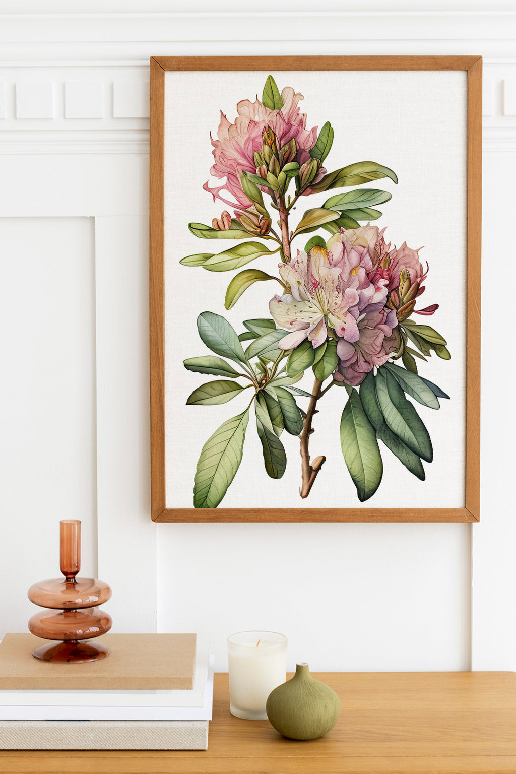 a framed botanical illustration of a pink and white flower on a white wall, with a wooden shelf below it holding a stack of books, a small green vase, and a white candle.