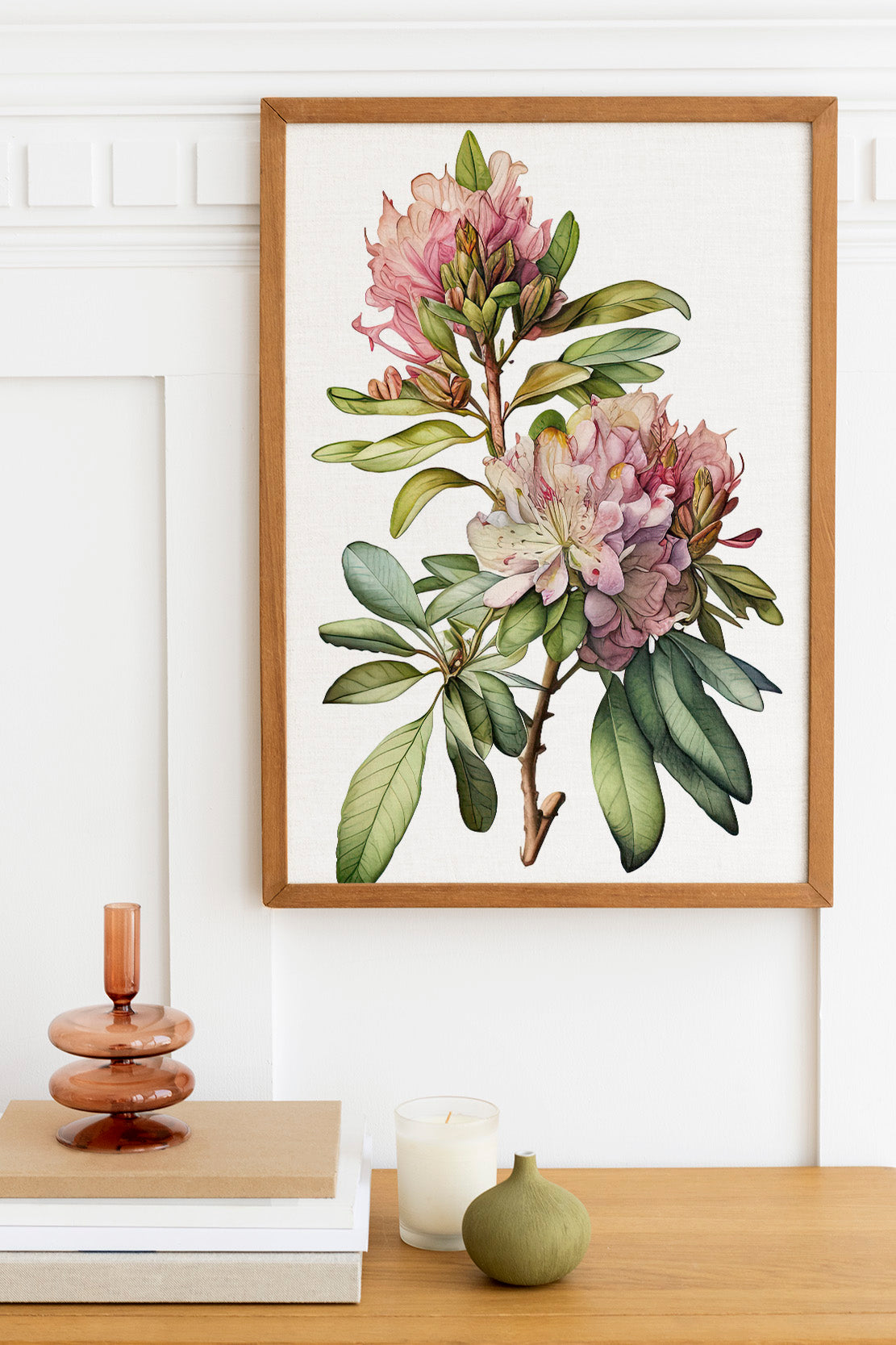 a framed botanical illustration of a pink and white flower on a white wall, with a wooden shelf below it holding a stack of books, a small green vase, and a white candle.
