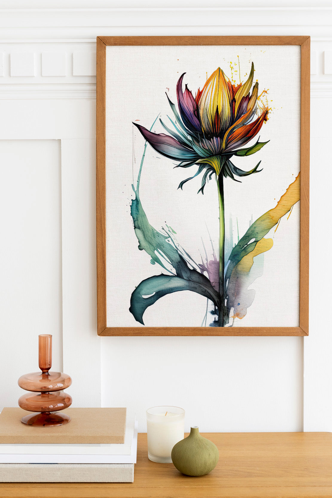 a framed painting of a colorful flower hanging on a wall above a wooden table with various decorative items, including a candle, a stack of books, and a small green vase.