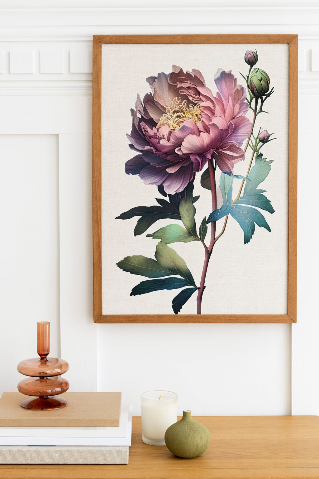 a framed painting of a large, vibrant pink peony flower hanging on a wall above a wooden table. The table also has a stack of books, a small green vase, and a white candle.