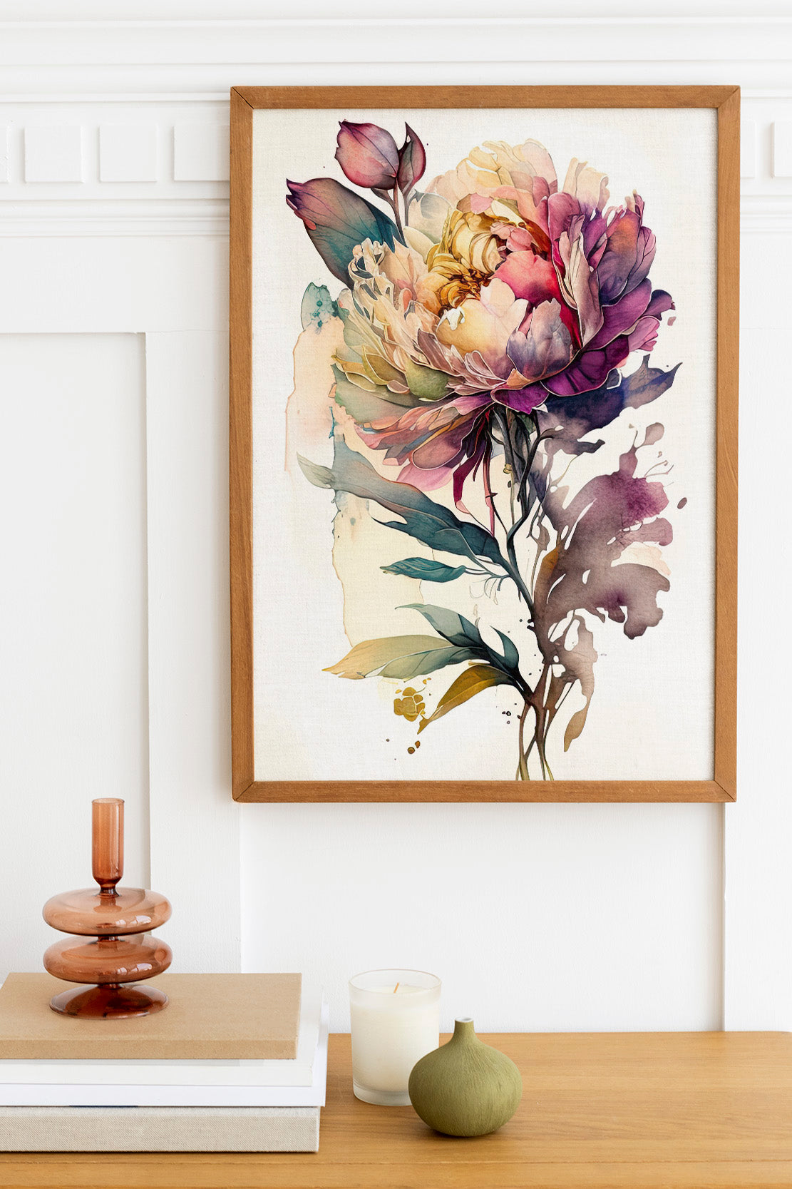 a framed watercolor painting of a large, colorful flower with purple, yellow, and green hues. The painting is hanging on a white wall above a wooden table with a stack of books and a small green vase.