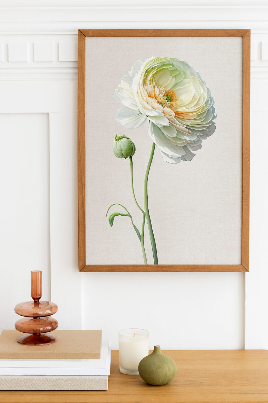 a wooden table with a framed painting of a large white and yellow flower hanging on the wall above it. On the table, there is a stack of books, a small green vase, and a white candle.