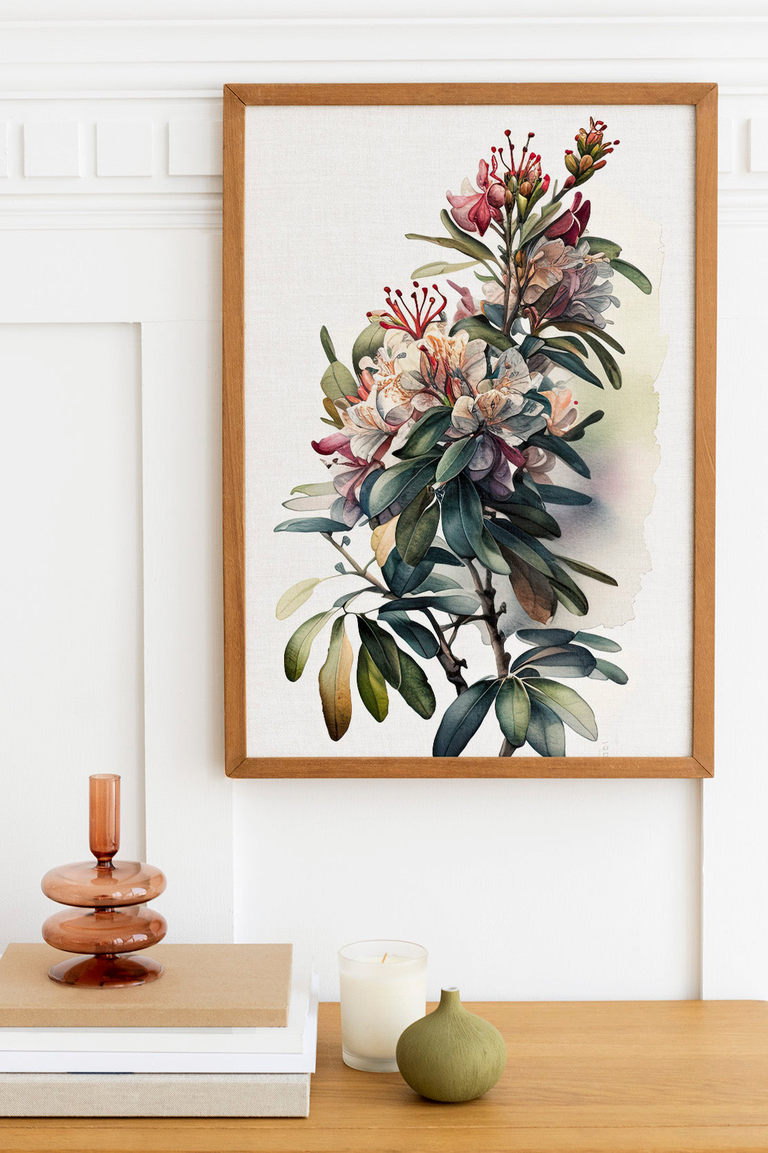a framed painting of a lush, colorful floral arrangement hanging on a white wall above a wooden table. The table also holds a small vase, a candle, and a stack of books.