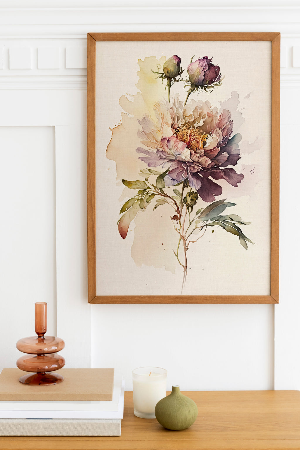 a framed watercolor painting of a large, colorful flower on a wall, with a wooden shelf below it holding a stack of books, a small green vase, and a white candle.