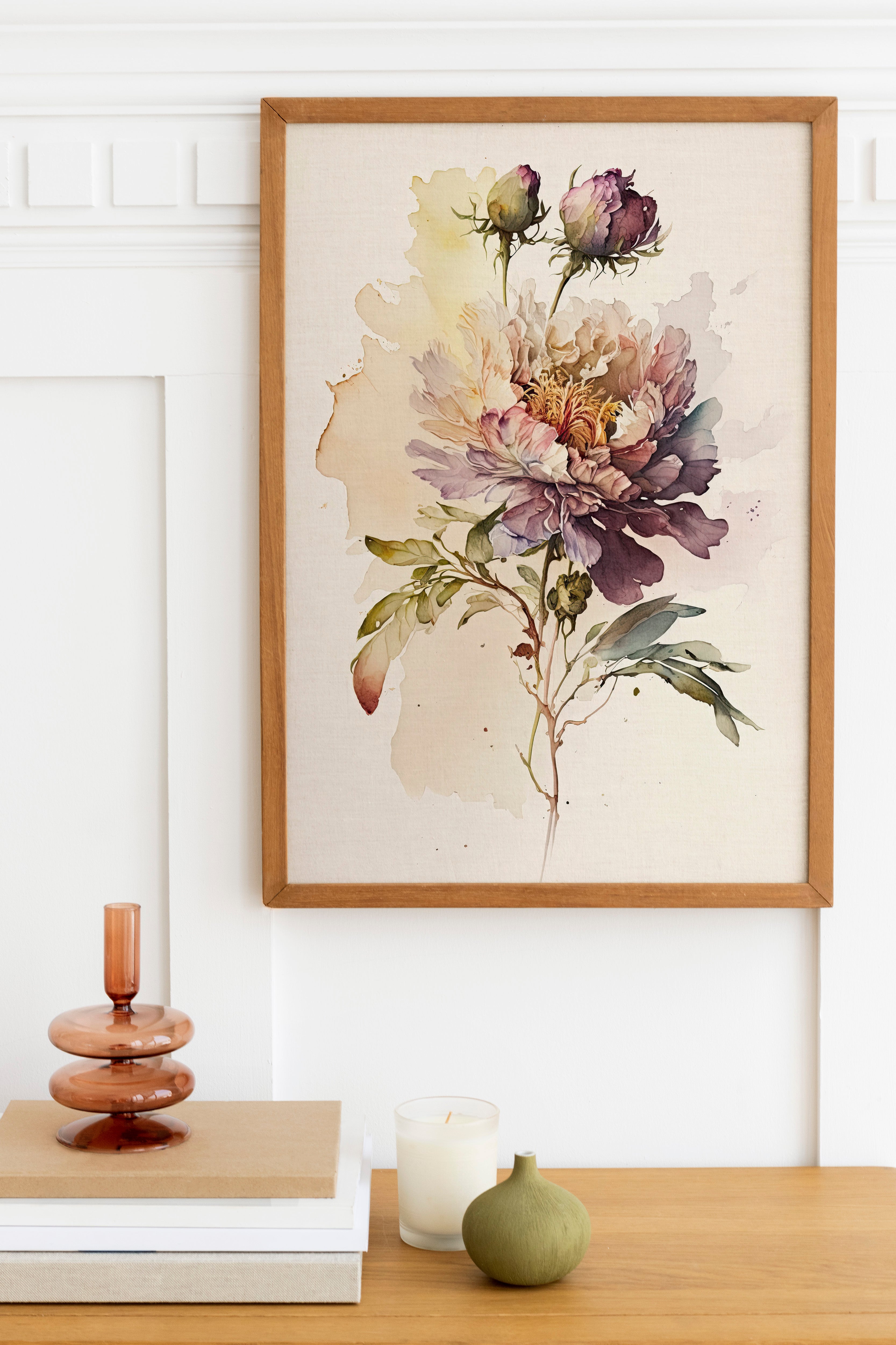a framed watercolor painting of a large, colorful flower on a wall, with a wooden shelf below it holding a stack of books, a small green vase, and a white candle.