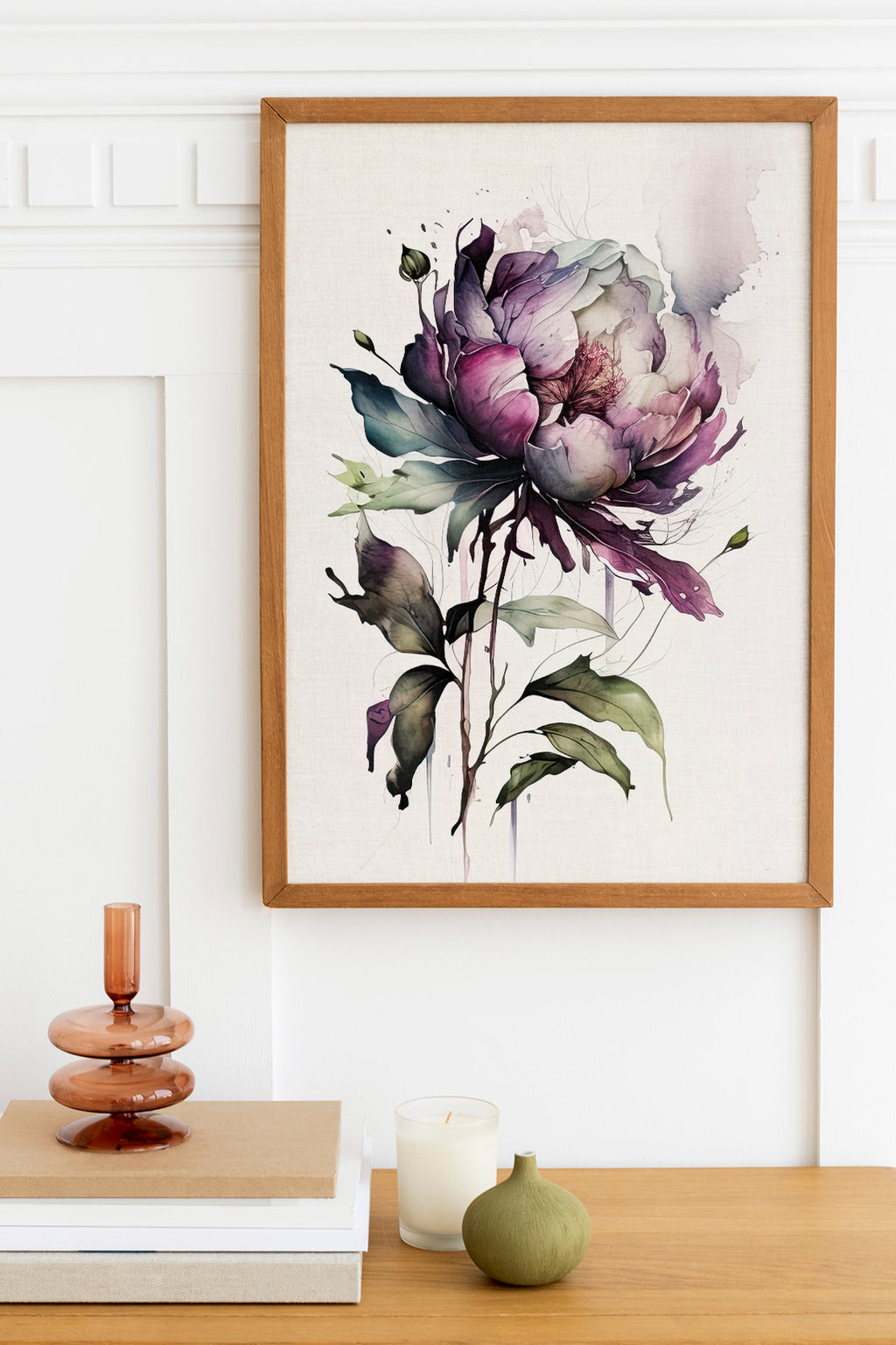 a framed watercolor painting of a large purple flower with green leaves, hanging on a white wall above a wooden table. On the table, there is a stack of books, a small green vase, and a white candle.