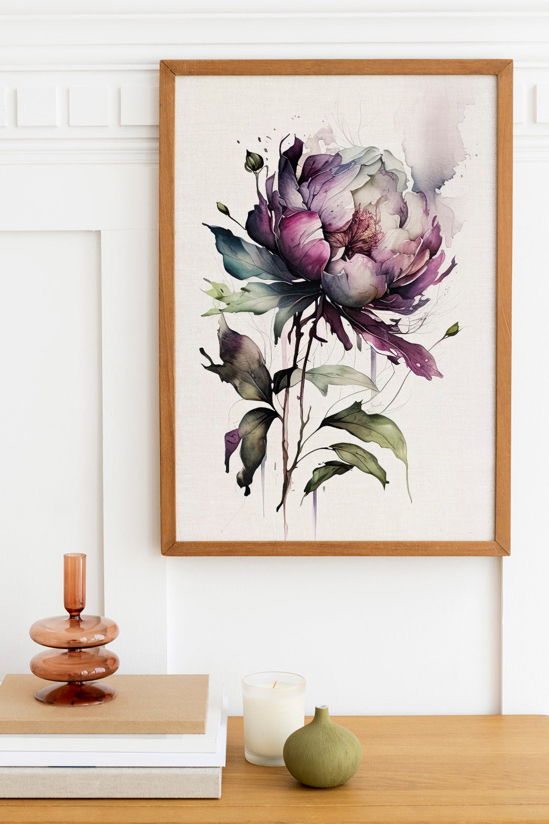 a framed watercolor painting of a large purple flower with green leaves, hanging on a white wall above a wooden table. On the table, there is a stack of books, a small green vase, and a white candle.