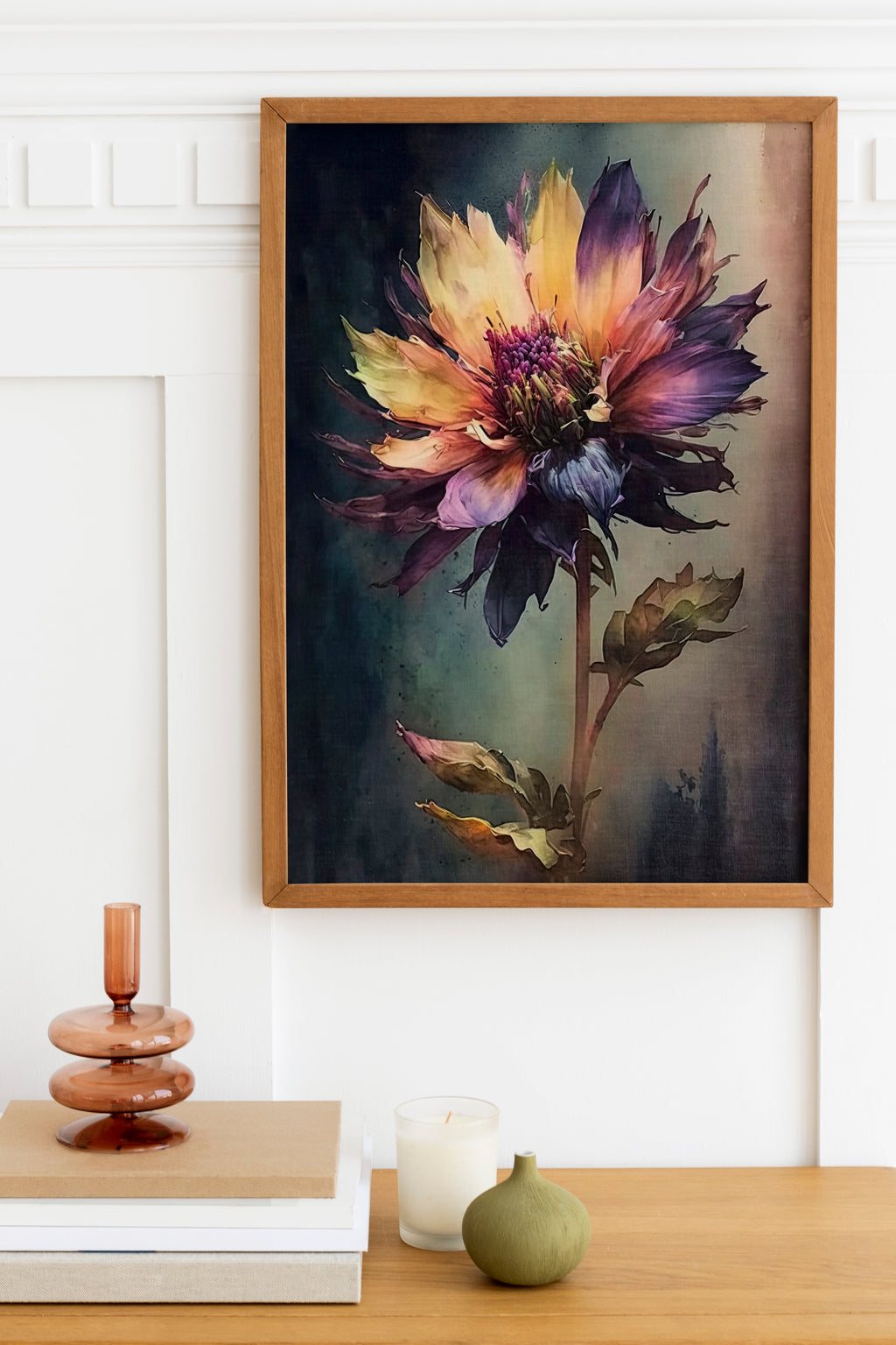 a framed painting of a vibrant, multi-colored flower hanging on a wall above a wooden table. The table has a stack of books, a small green vase, and a white candle.