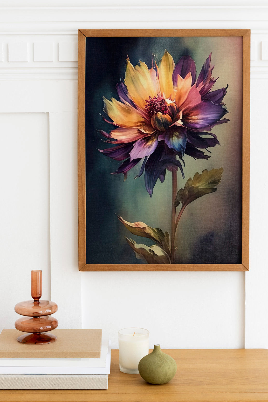 a framed painting of a vibrant, multi-colored flower hanging on a wall above a wooden table. On the table, there is a stack of books, a small green vase, and a white candle.