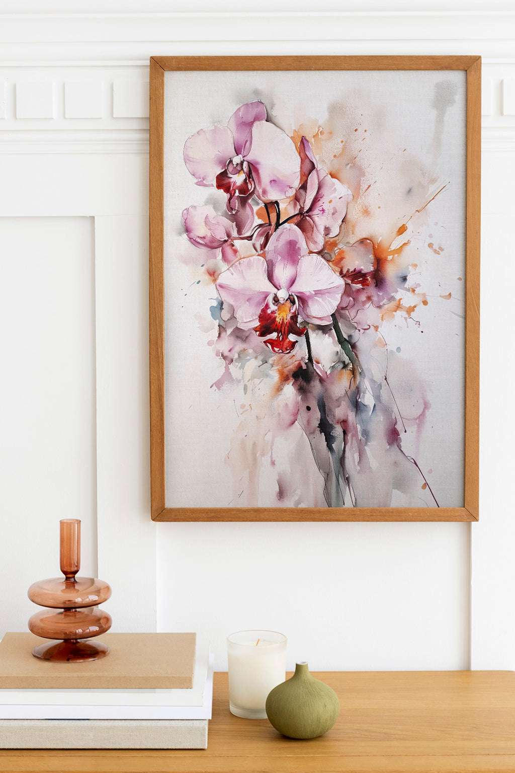 a framed painting of a bouquet of pink orchids hanging on a wall above a wooden table with various decorative items, including a candle, a small green vase, and a stack of books.