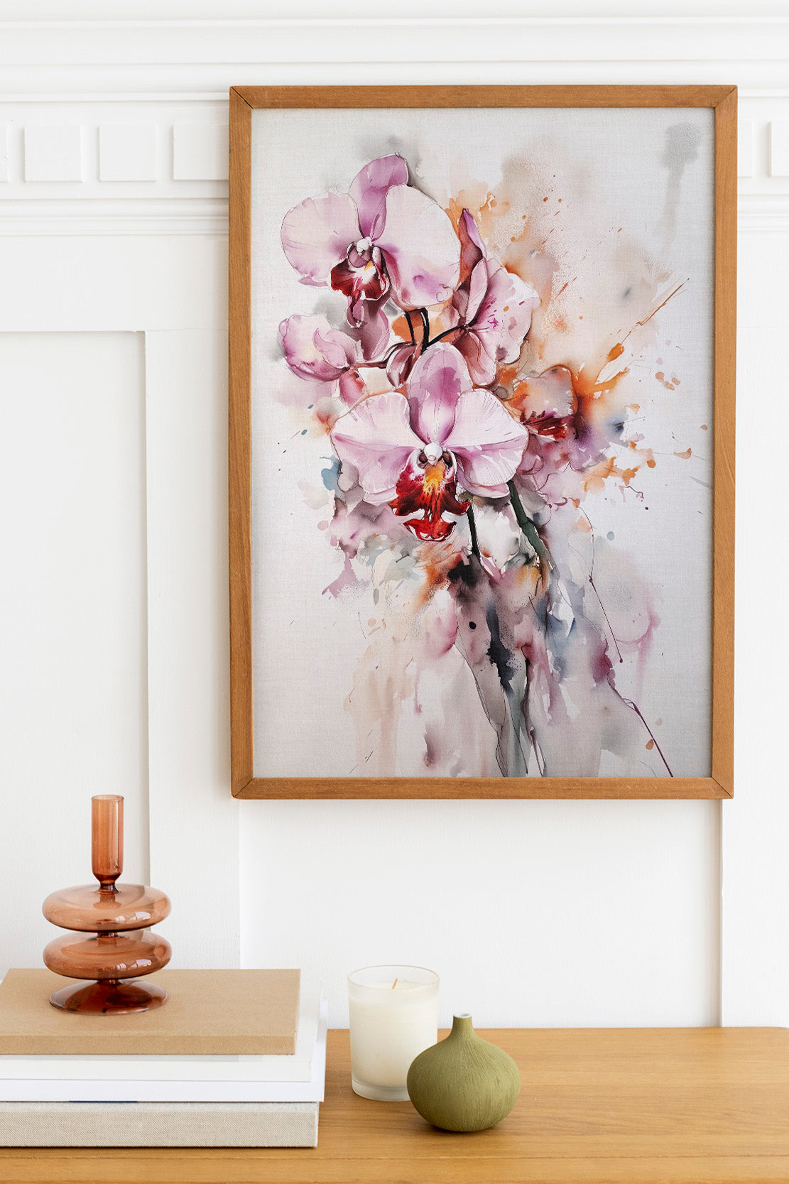 a framed painting of a bouquet of pink orchids hanging on a wall above a wooden table with various decorative items, including a candle, a small green vase, and a stack of books.