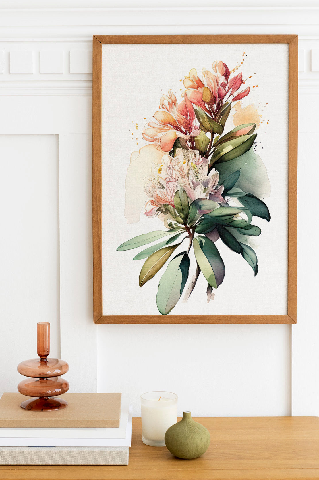 a framed watercolor painting of a vibrant floral arrangement hanging on a white wall above a wooden table. The table also holds a stack of books, a small green vase, and a white candle.