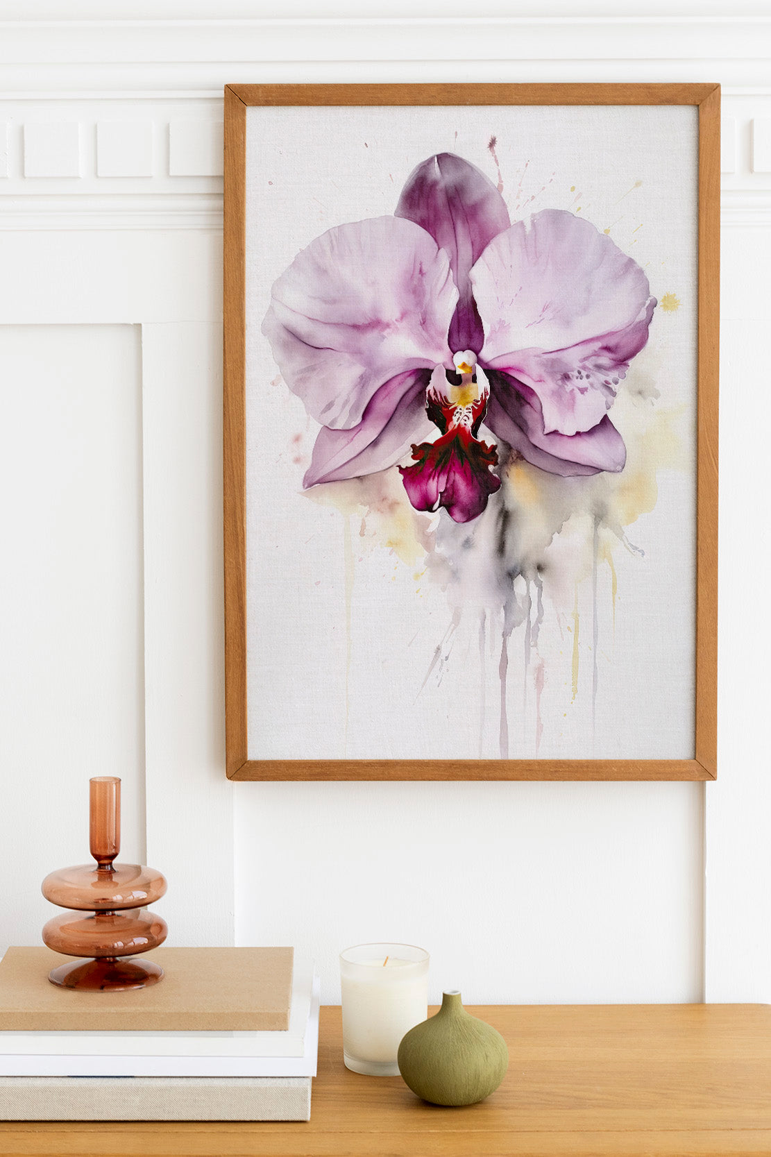 a framed watercolor painting of a purple orchid flower hanging on a white wall above a wooden table. On the table, there is a stack of books, a small green vase, and a white candle.