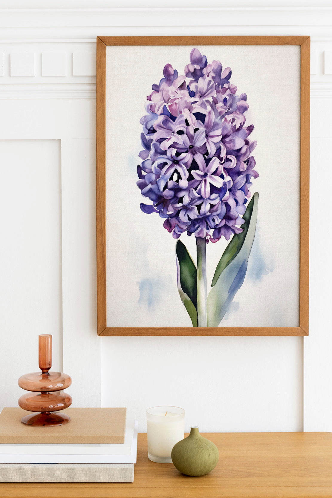a framed painting of a purple hyacinth flower hanging on a white wall above a wooden table. On the table, there is a stack of books, a small green vase, and a white candle.