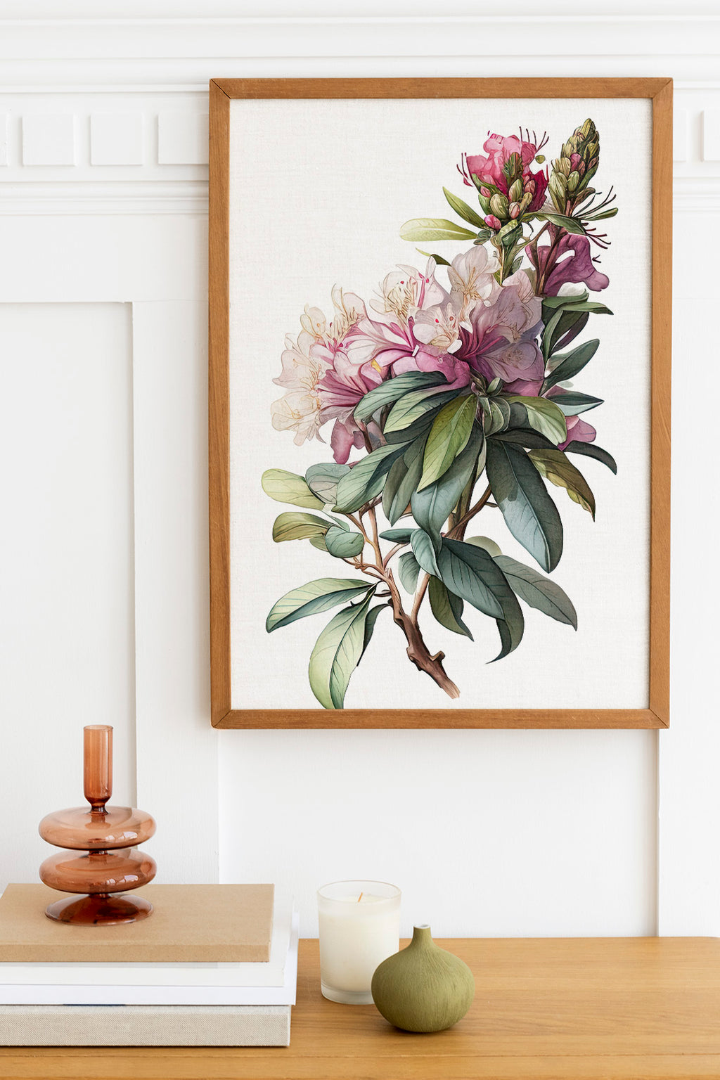 a framed botanical illustration of a flower with green leaves, hanging on a white wall above a wooden table. On the table, there is a stack of books, a small green vase, and a white candle.