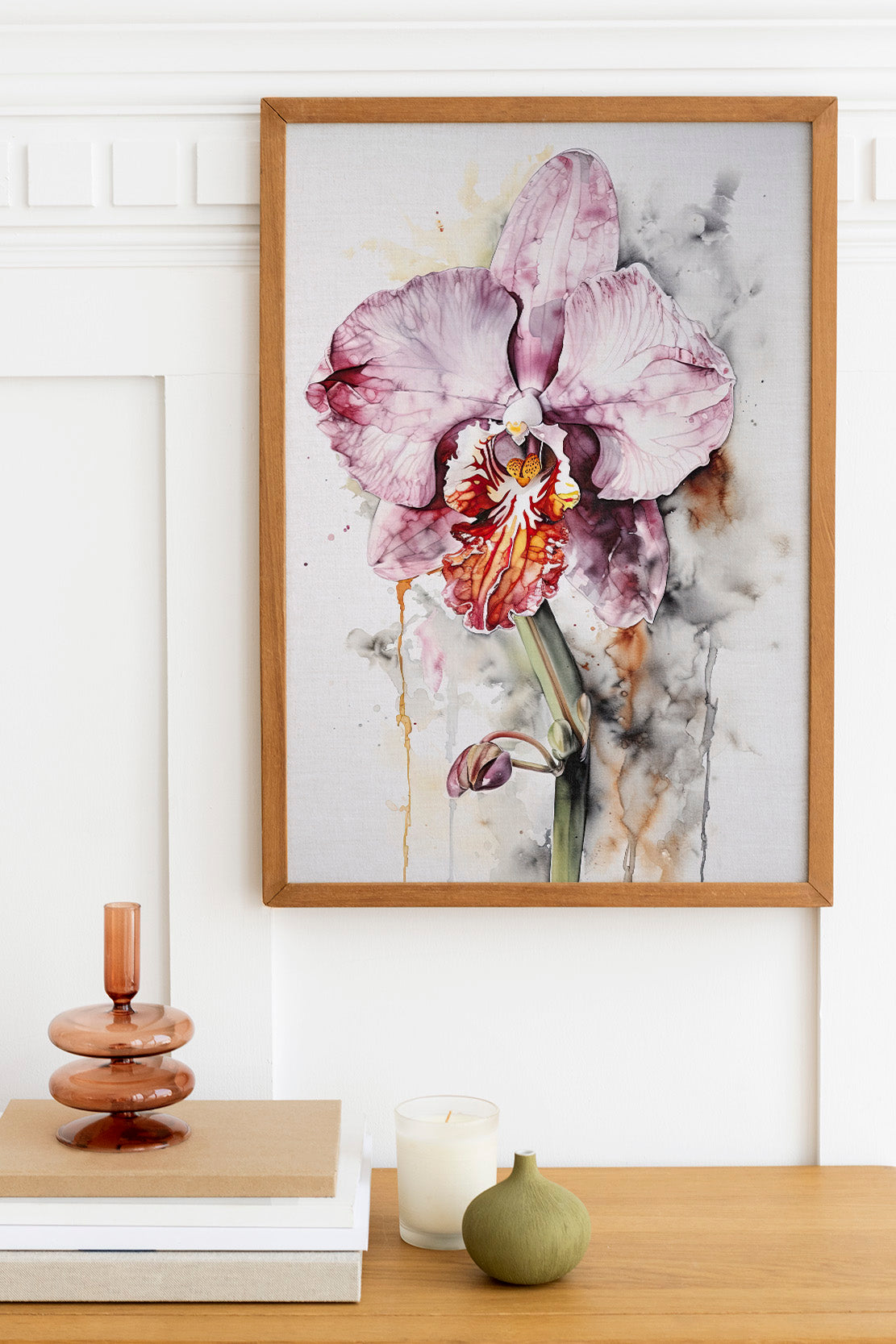 a framed painting of a purple orchid hanging on a wall above a wooden table with various decorative items, including a candle, a stack of books, and a small green vase.