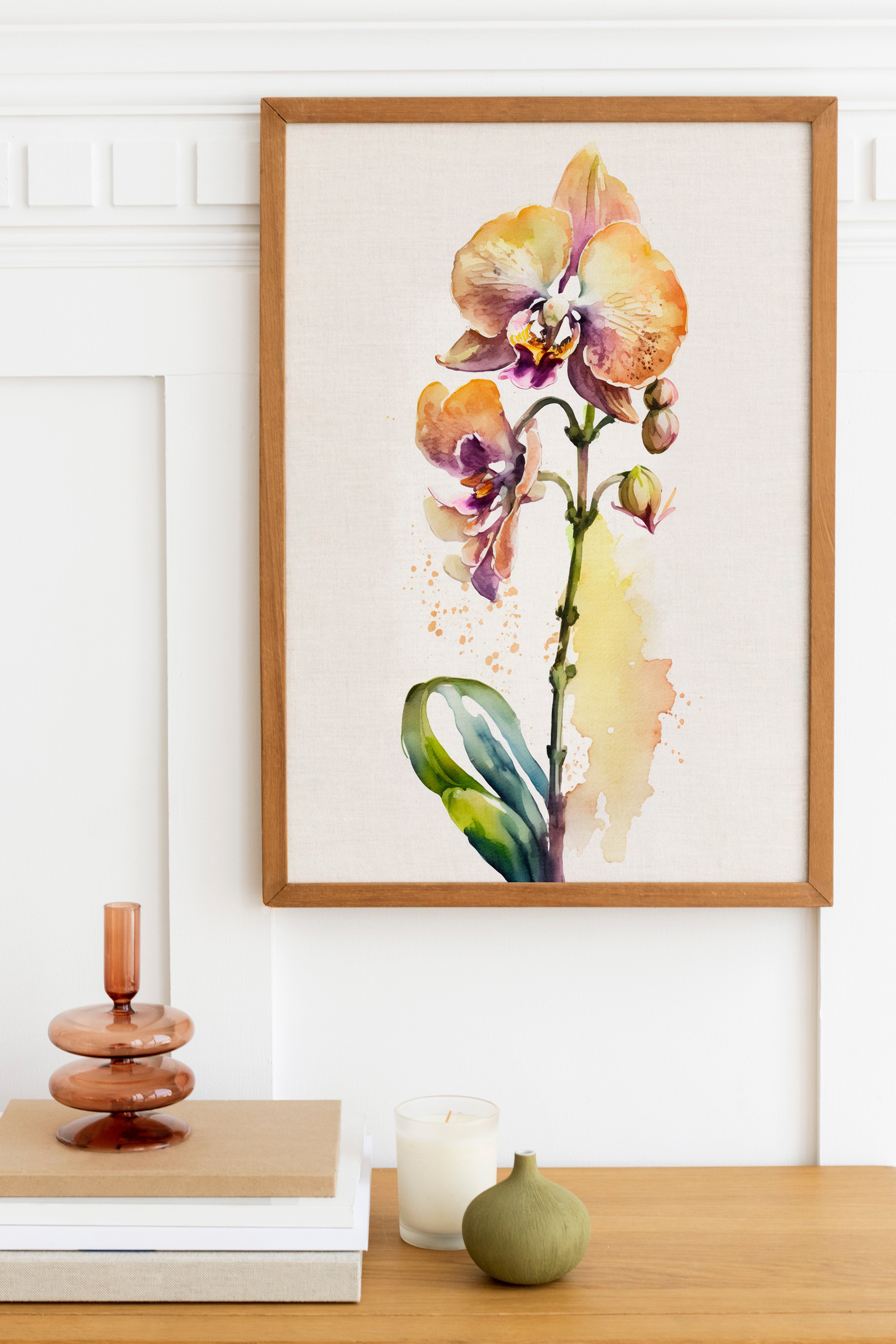 a framed watercolor painting of an orchid flower hanging on a wall above a wooden table with various decorative items, including a candle, a stack of books, and a small green vase.