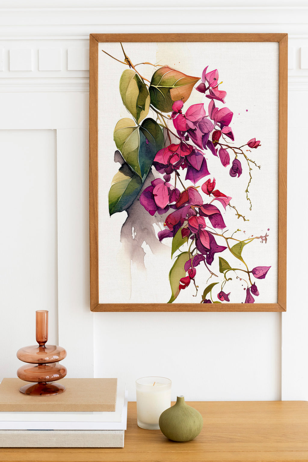 a framed watercolor painting of purple flowers hanging on a wall above a wooden table with various decorative items, including a candle, a stack of books, and a small green vase.