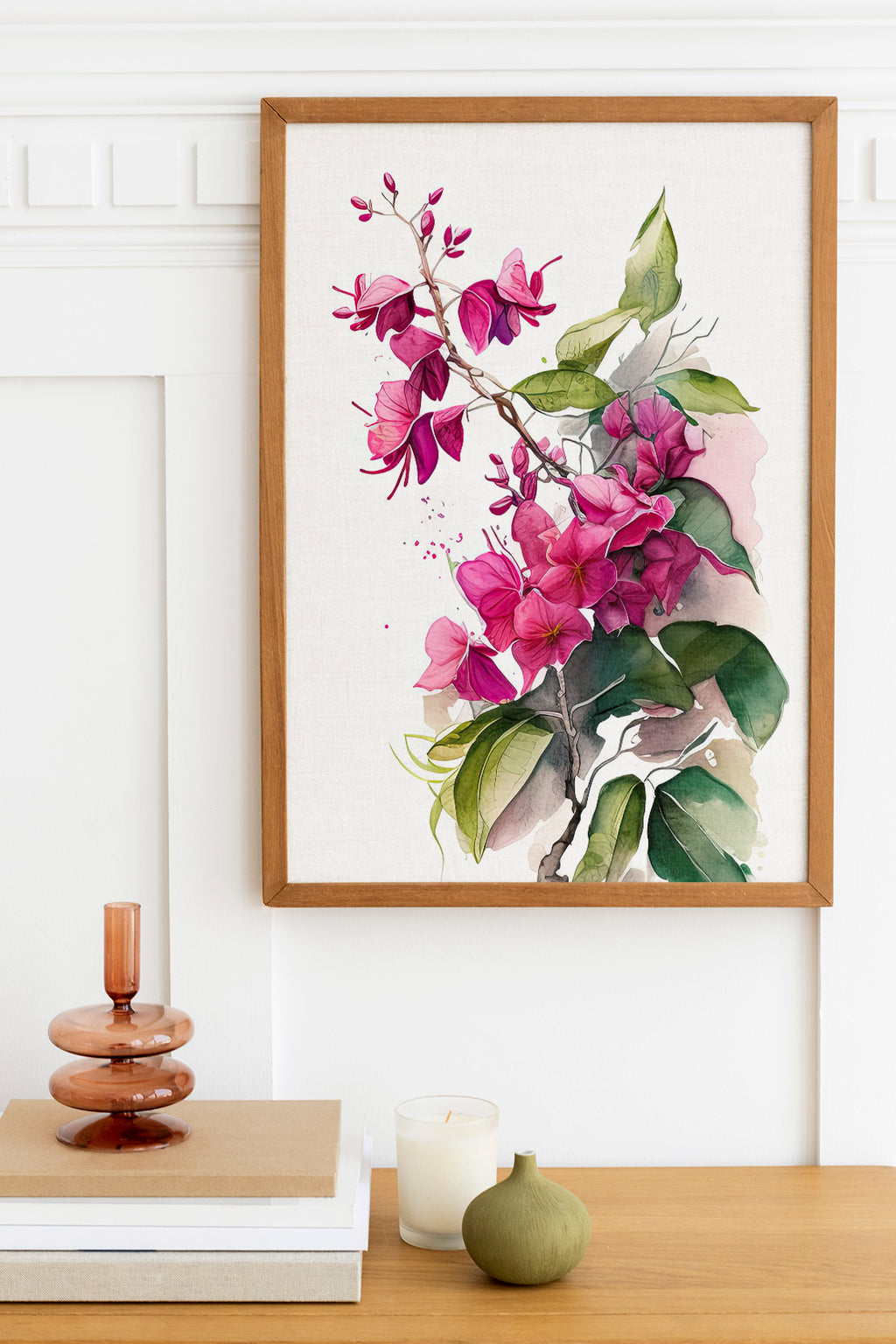 a framed painting of a branch with pink flowers and green leaves hanging on a white wall above a wooden table. On the table, there is a stack of books, a small green vase, and a white candle.