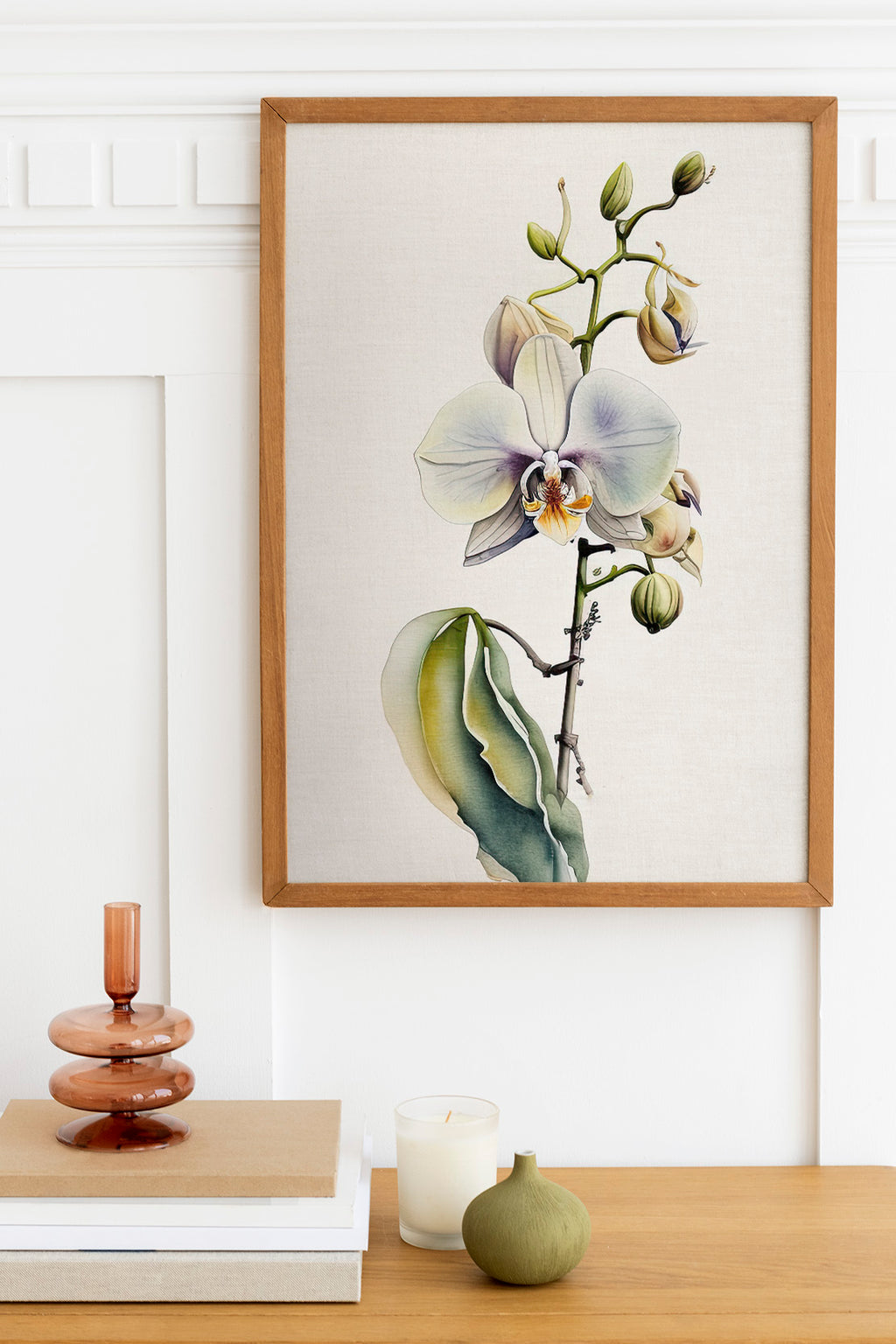 a framed painting of a white orchid flower hanging on a wall above a wooden table with various decorative items, including a candle, a small green vase, and a stack of books.