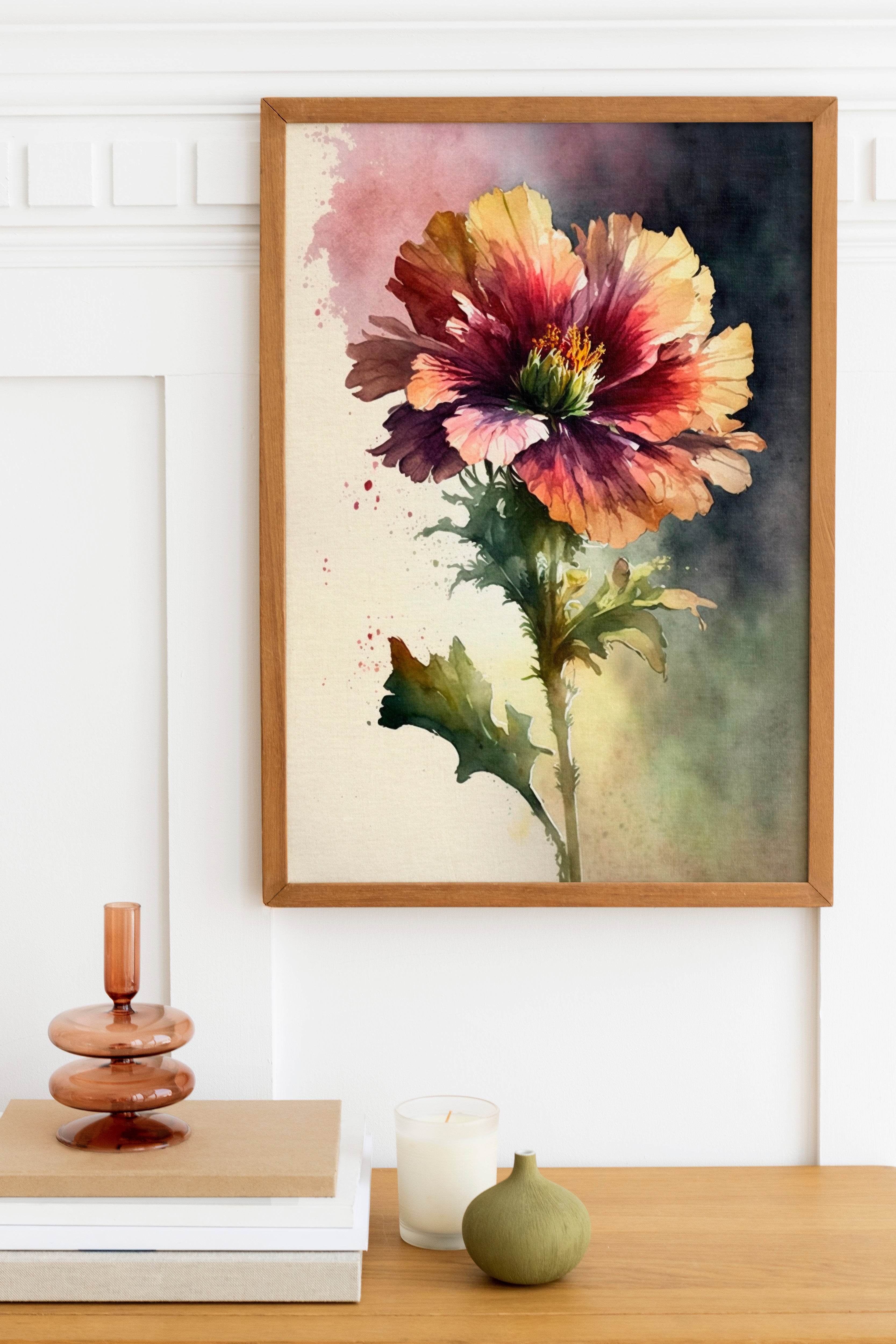 a framed painting of a vibrant, colorful flower hanging on a wall above a wooden table. The table also holds a stack of books, a small green vase, and a white candle.