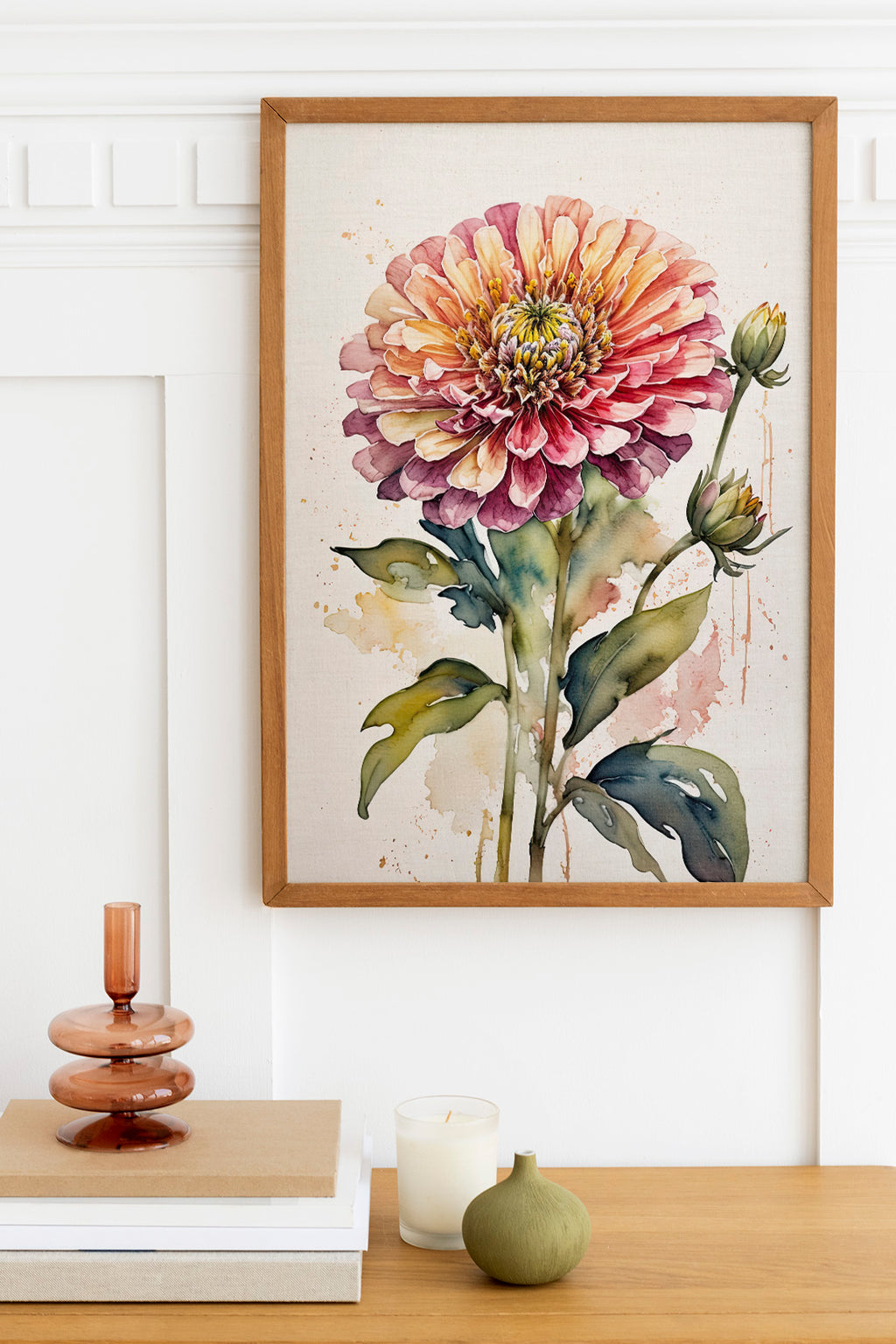 a framed watercolor painting of a large, vibrant flower with a yellow center and pink petals, hanging on a white wall above a wooden table. The table also has a stack of books, a small green vase, and a white candle.