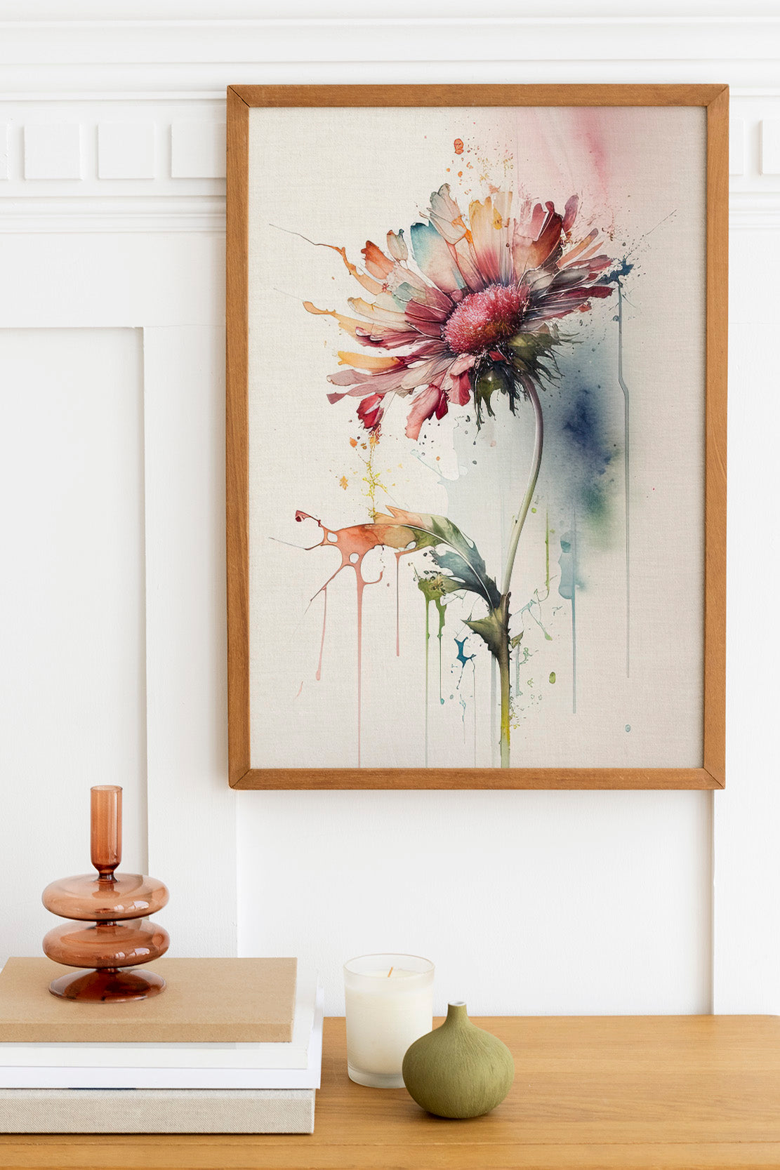 a framed watercolor painting of a vibrant flower hanging on a wall above a wooden table. The table has a stack of books, a small green vase, and a white candle.