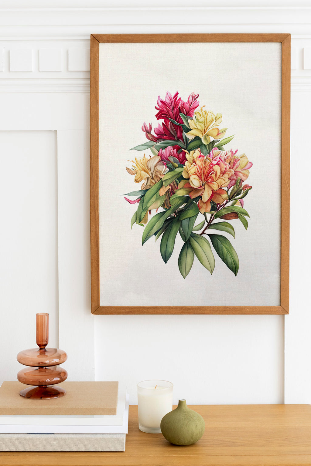 a framed painting of a vibrant bouquet of flowers hanging on a wall above a wooden table. The table also holds a stack of books, a small green vase, and a white candle.