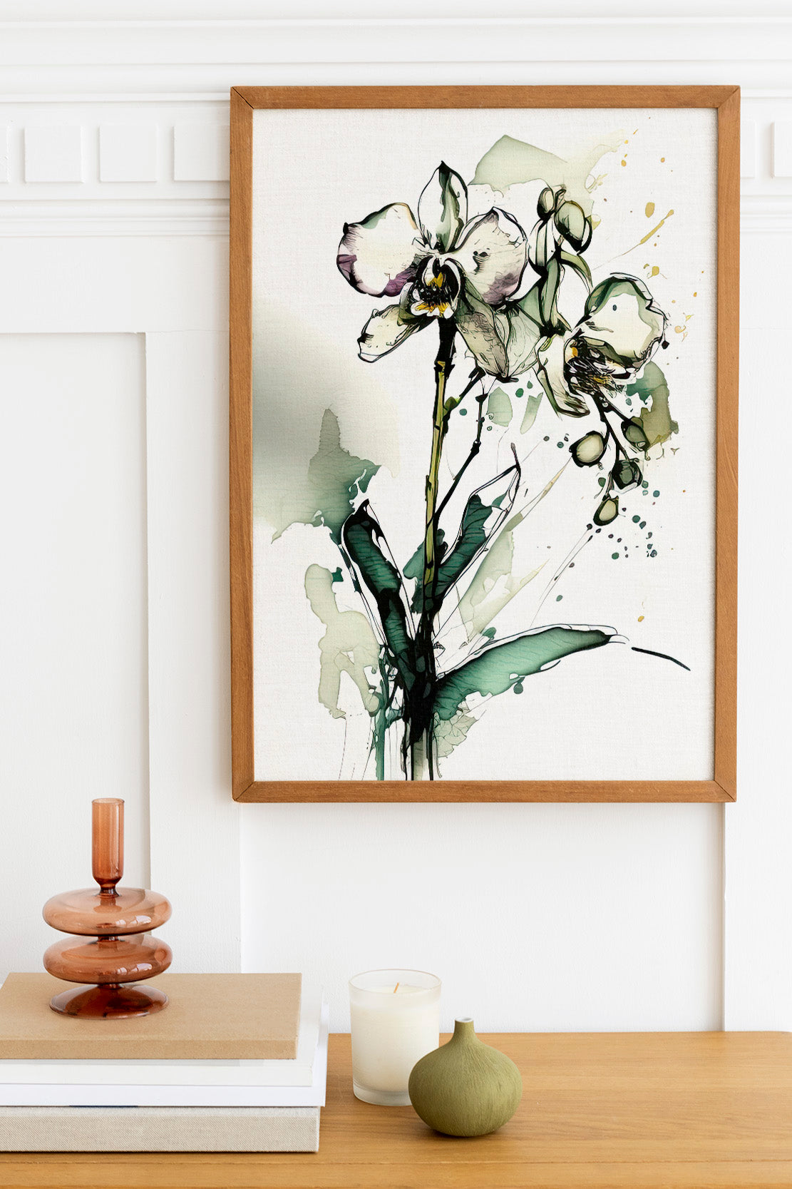 a framed painting of a white orchid flower with green leaves, hanging on a white wall above a wooden table. On the table, there is a stack of books, a small green vase, and a white candle.