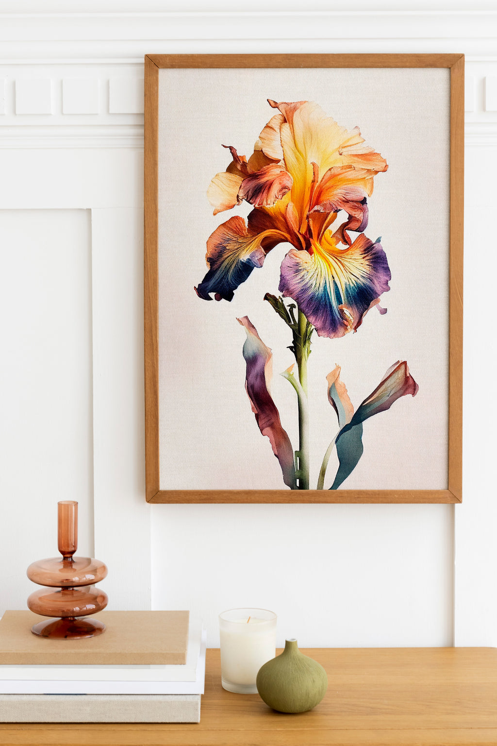 a framed painting of a vibrant, multi-colored iris flower hanging on a wall above a wooden table. The table also has a stack of books, a small green vase, and a white candle.