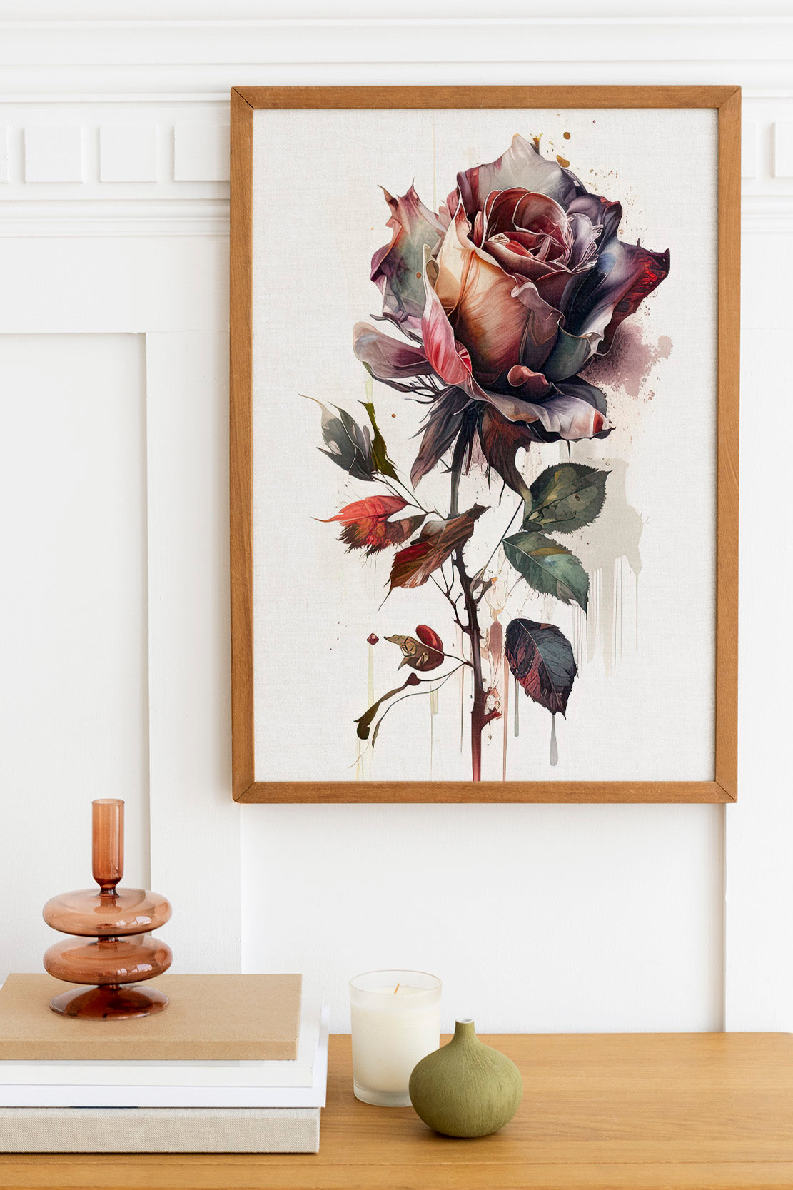 a framed painting of a large, colorful rose hanging on a wall above a wooden table with various decorative items, including a candle, a stack of books, and a small green vase.