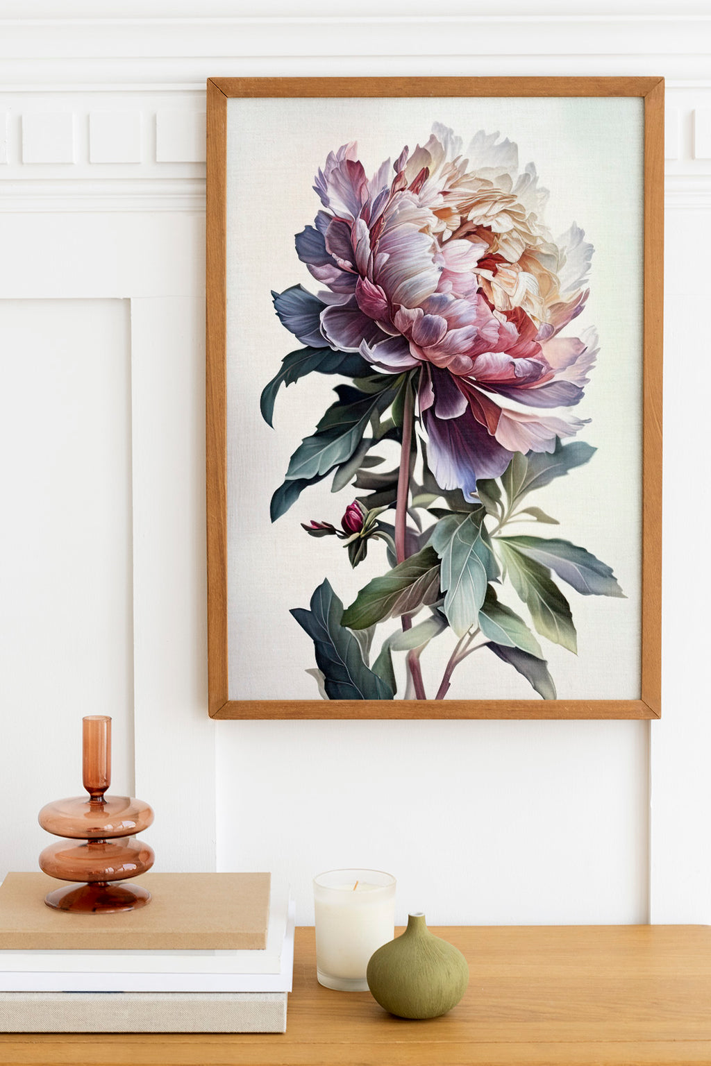 a framed painting of a large, colorful flower hanging on a wall above a wooden table. On the table, there is a stack of books, a small green vase, and a white candle.