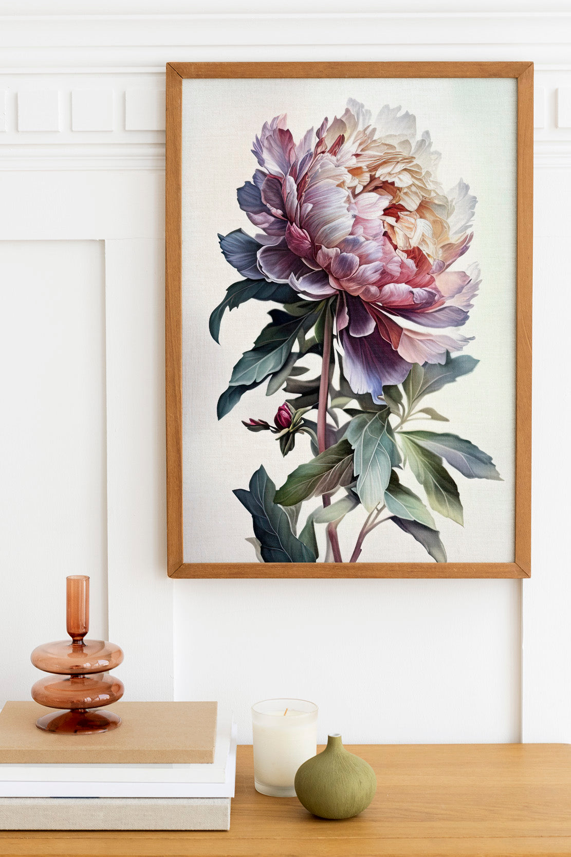 a framed painting of a large, colorful flower hanging on a wall above a wooden table. On the table, there is a stack of books, a small green vase, and a white candle.