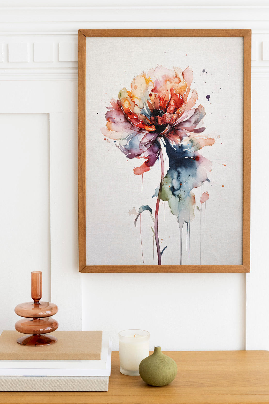 a wooden table with a framed watercolor painting of a colorful flower hanging on the wall above it. The table also has a stack of books, a small green vase, and a white candle.