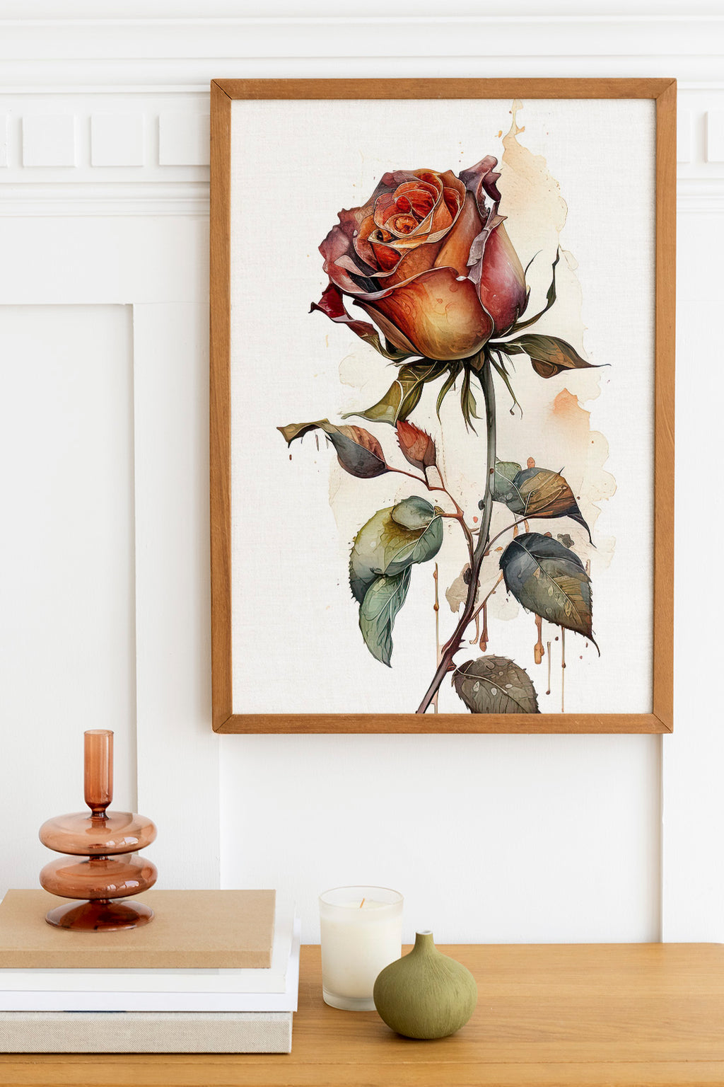 a framed painting of a large, vibrant rose hanging on a wall above a wooden table. The table also has a stack of books, a small green vase, and a white candle.