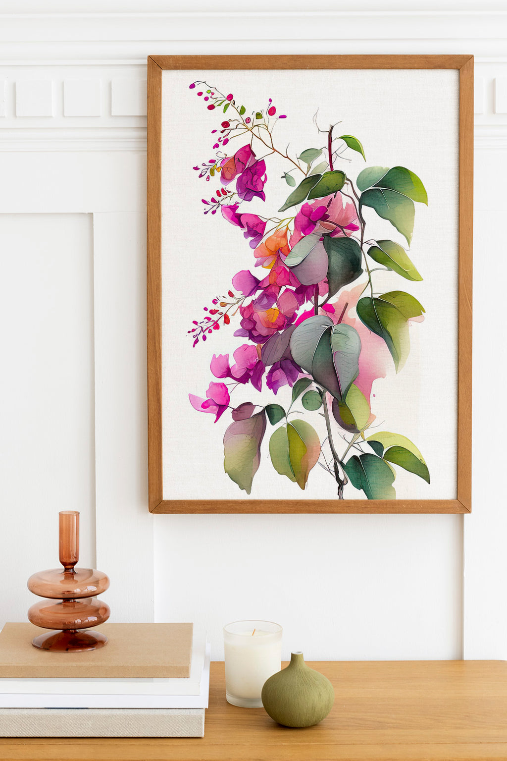 a framed painting of a vibrant floral arrangement hanging on a white wall above a wooden table. The painting is surrounded by various decorative items, including a stack of books, a small green vase, and a white candle.