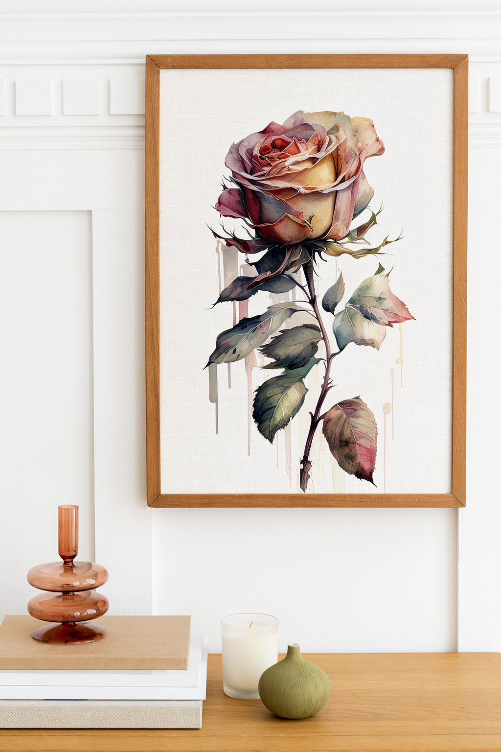 a framed painting of a large, vibrant rose hanging on a wall above a wooden table. The table has a stack of books, a small green vase, and a white candle.