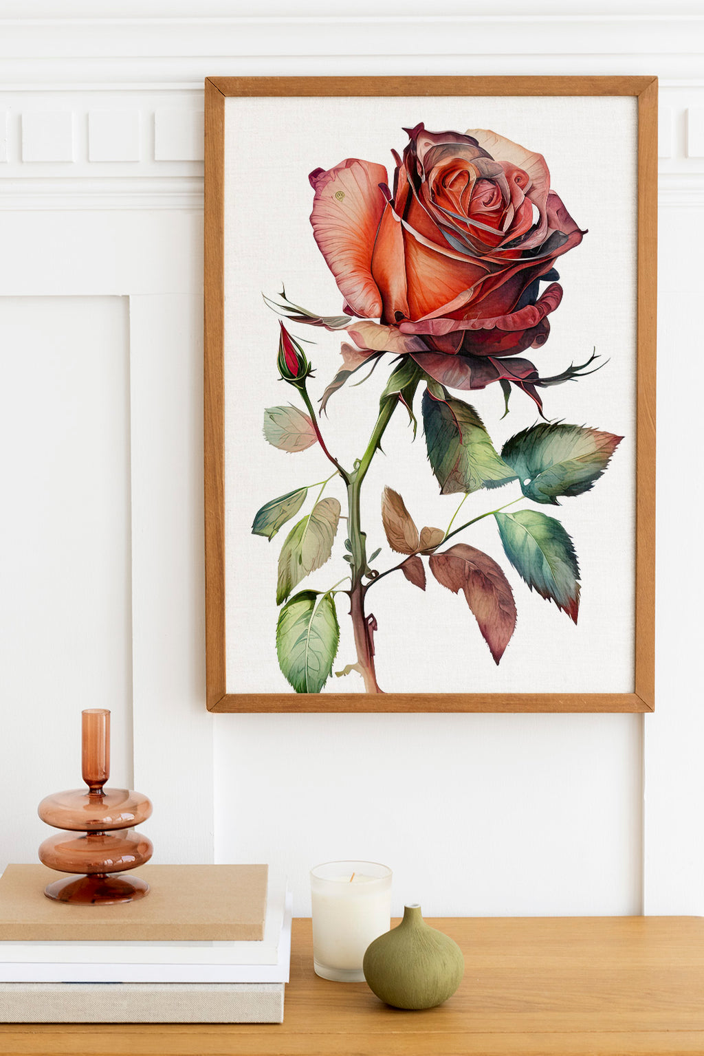 a framed painting of a large, vibrant red rose with green leaves, hanging on a white wall above a wooden table with various decorative items, including a candle, a small green vase, and a stack of books.