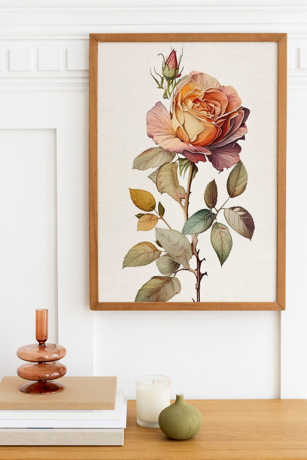 a framed print of a large, colorful rose on a wall, with a wooden shelf below it holding a stack of books, a small green vase, and a white candle.
