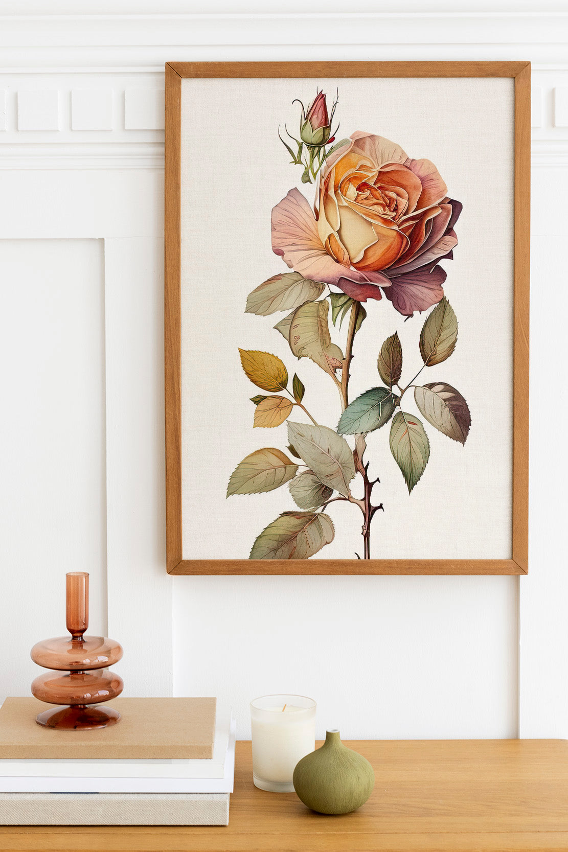 a framed print of a large, colorful rose on a wall, with a wooden shelf below it holding a stack of books, a small green vase, and a white candle.