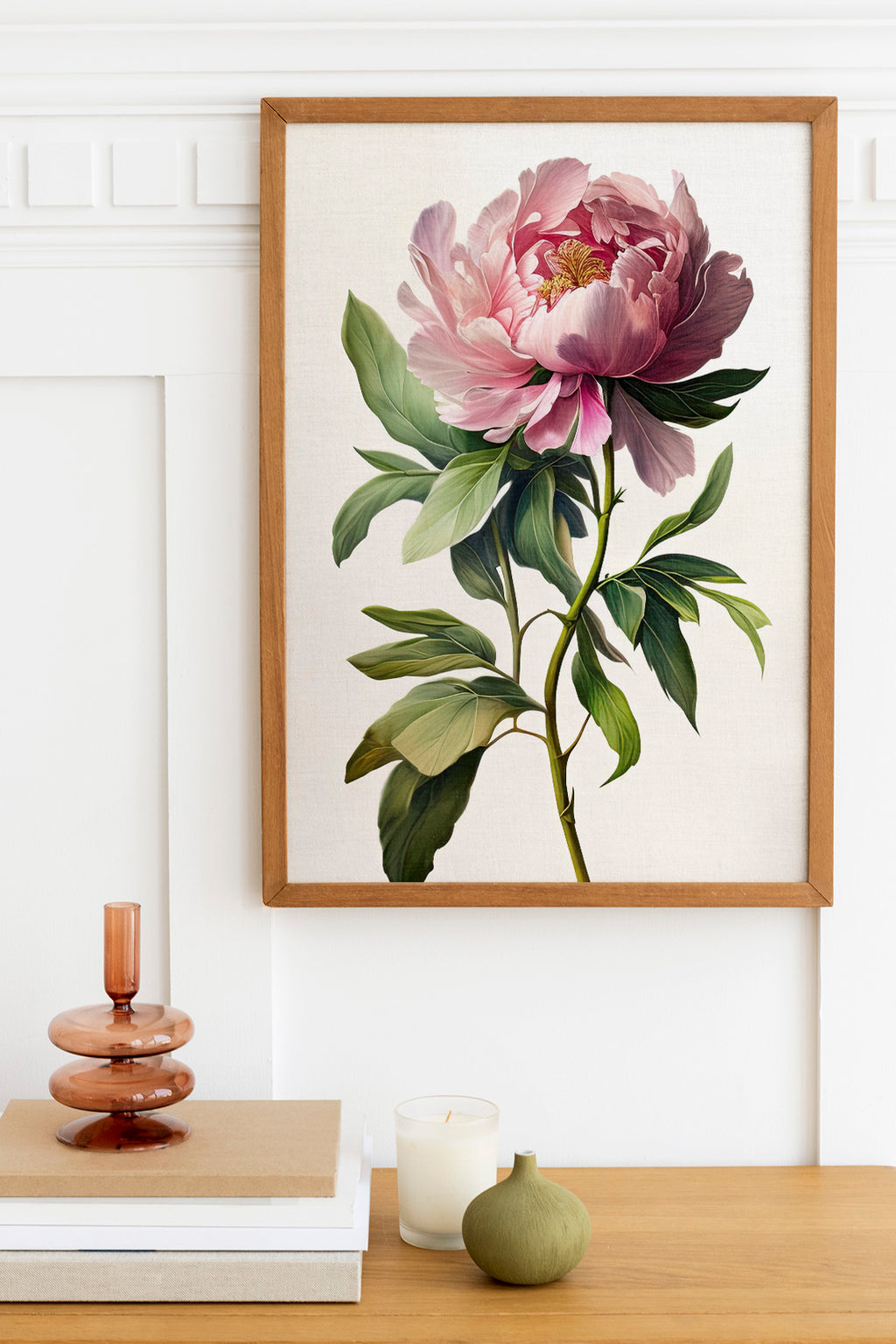 a framed painting of a large pink peony flower hanging on a white wall above a wooden table. On the table, there is a stack of books, a small green vase, and a white candle.