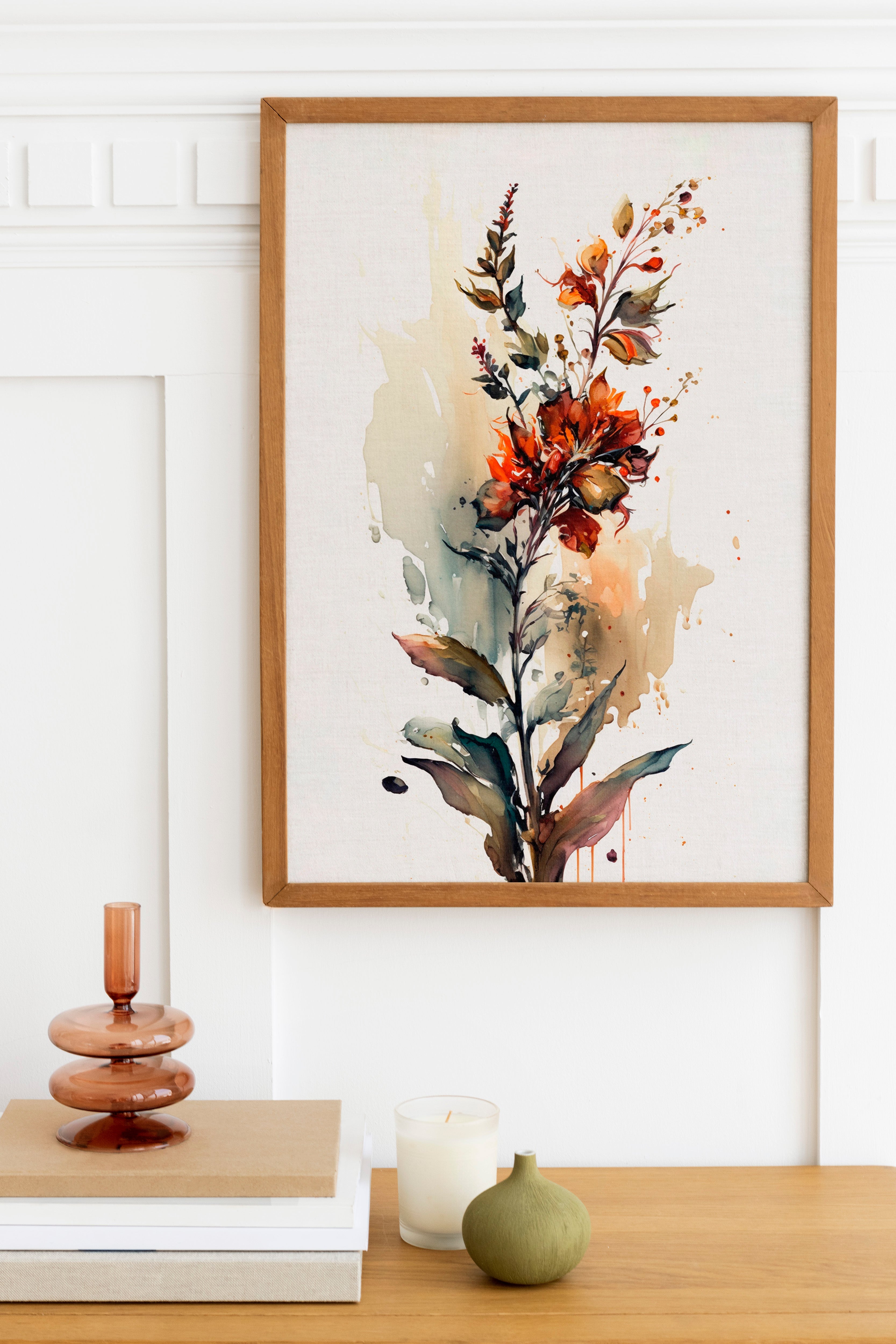a framed painting of a vibrant floral arrangement hanging on a wall above a wooden table. The table also holds a stack of books, a small green vase, and a white candle.