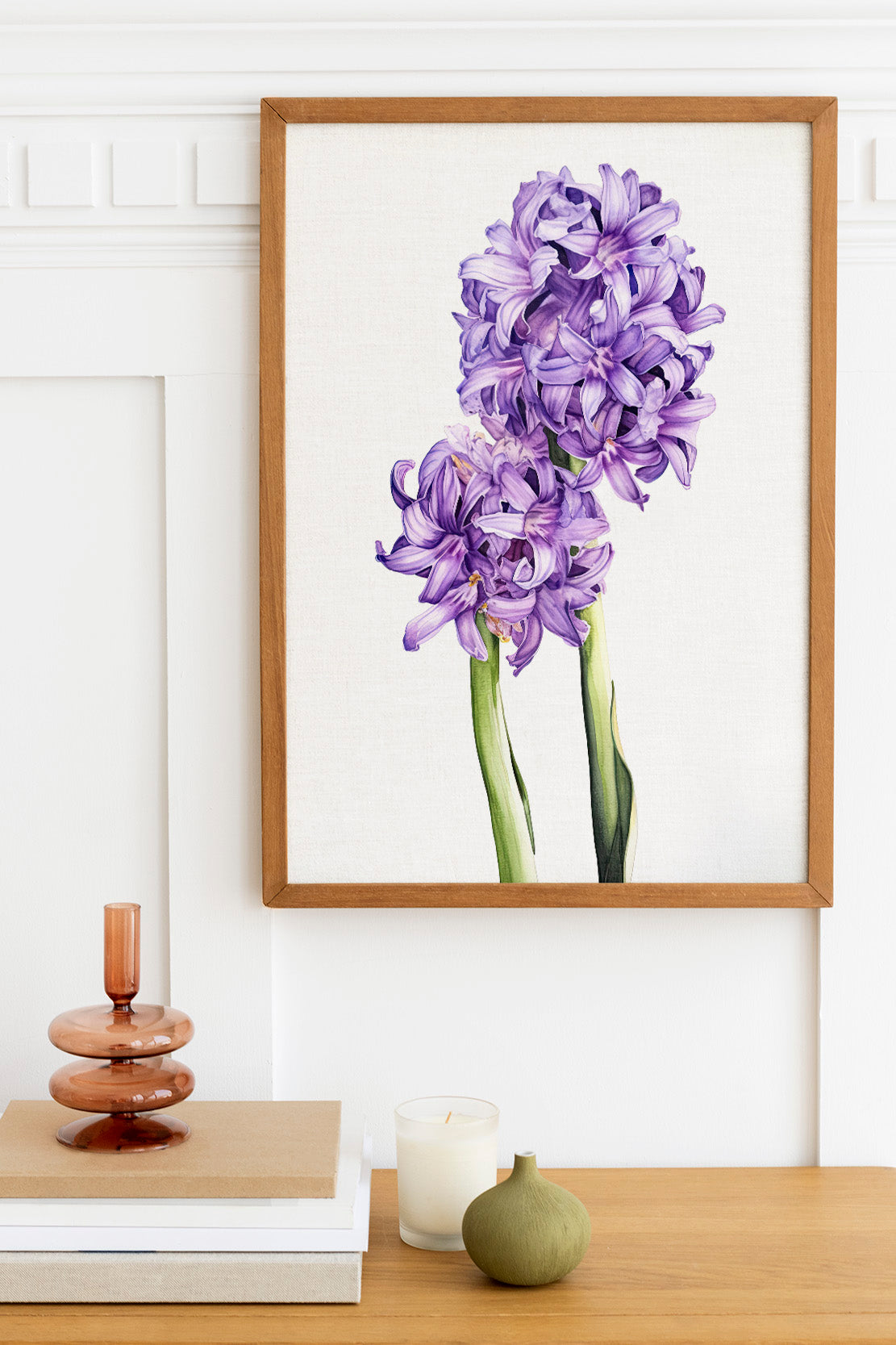 a framed painting of purple flowers hanging on a white wall above a wooden table. On the table, there is a stack of books, a small green vase, and a white candle.